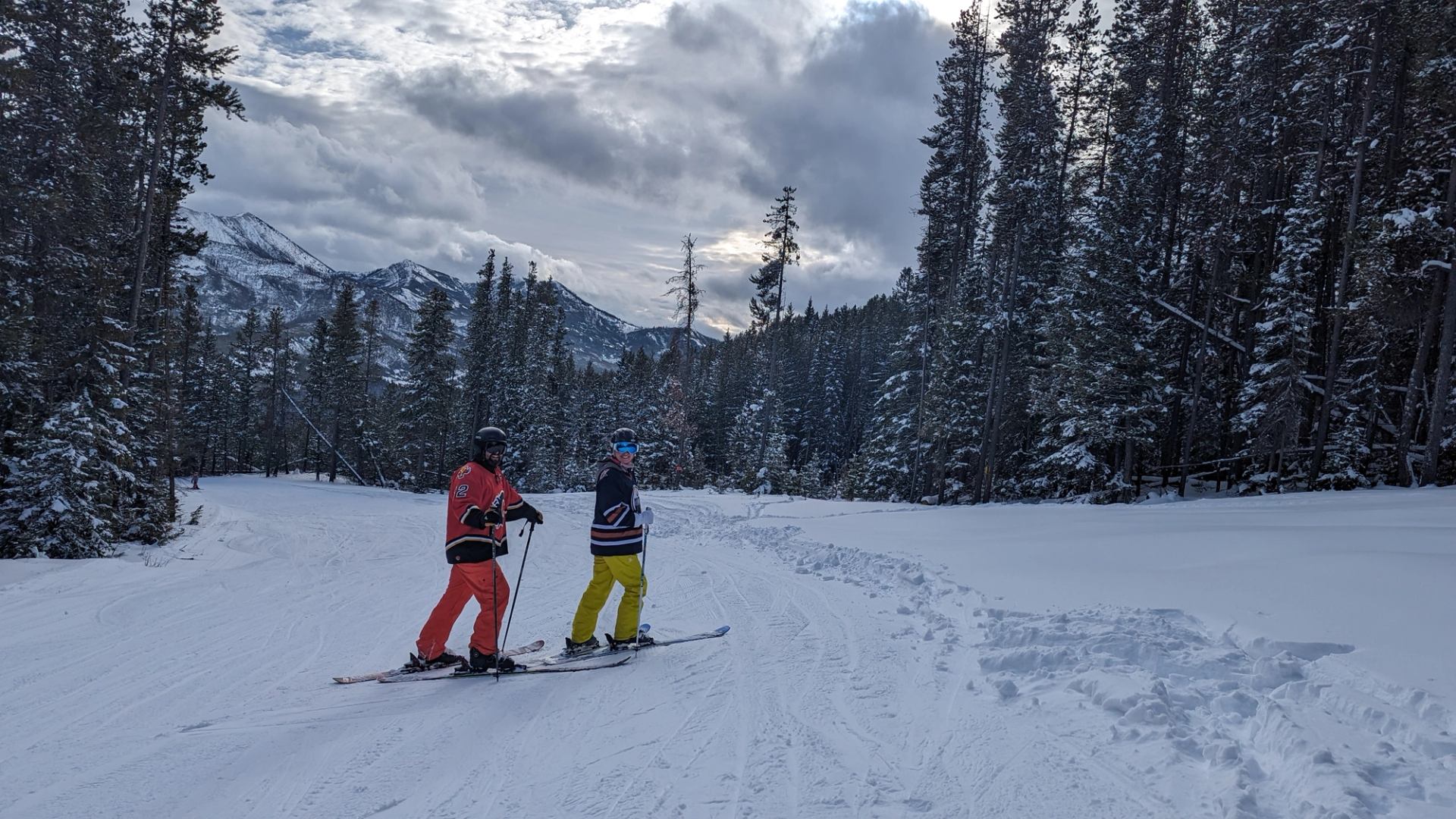 Skiers in hockey jerseys on snowy trail among pine trees with mountain backdrop.