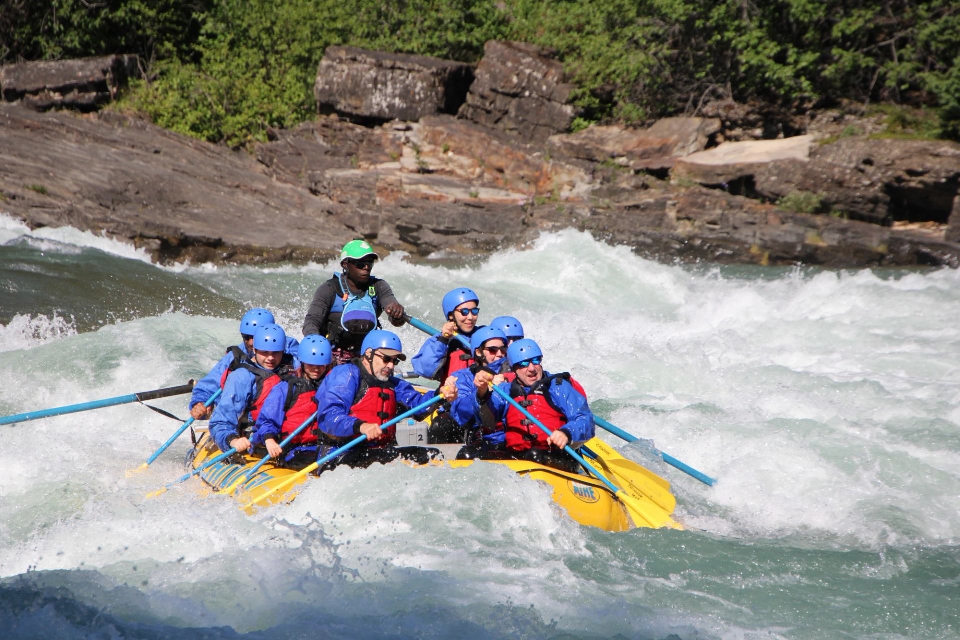 Group of rafters in helmets and lifejackets paddling through whitewater rapids.