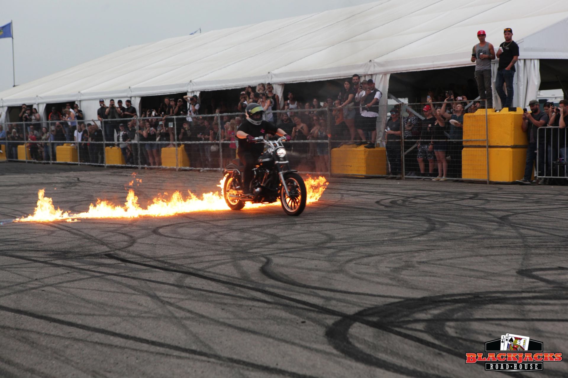 Motorcycle stunt rider performs with flames from bike and crowd behind metal barriers.