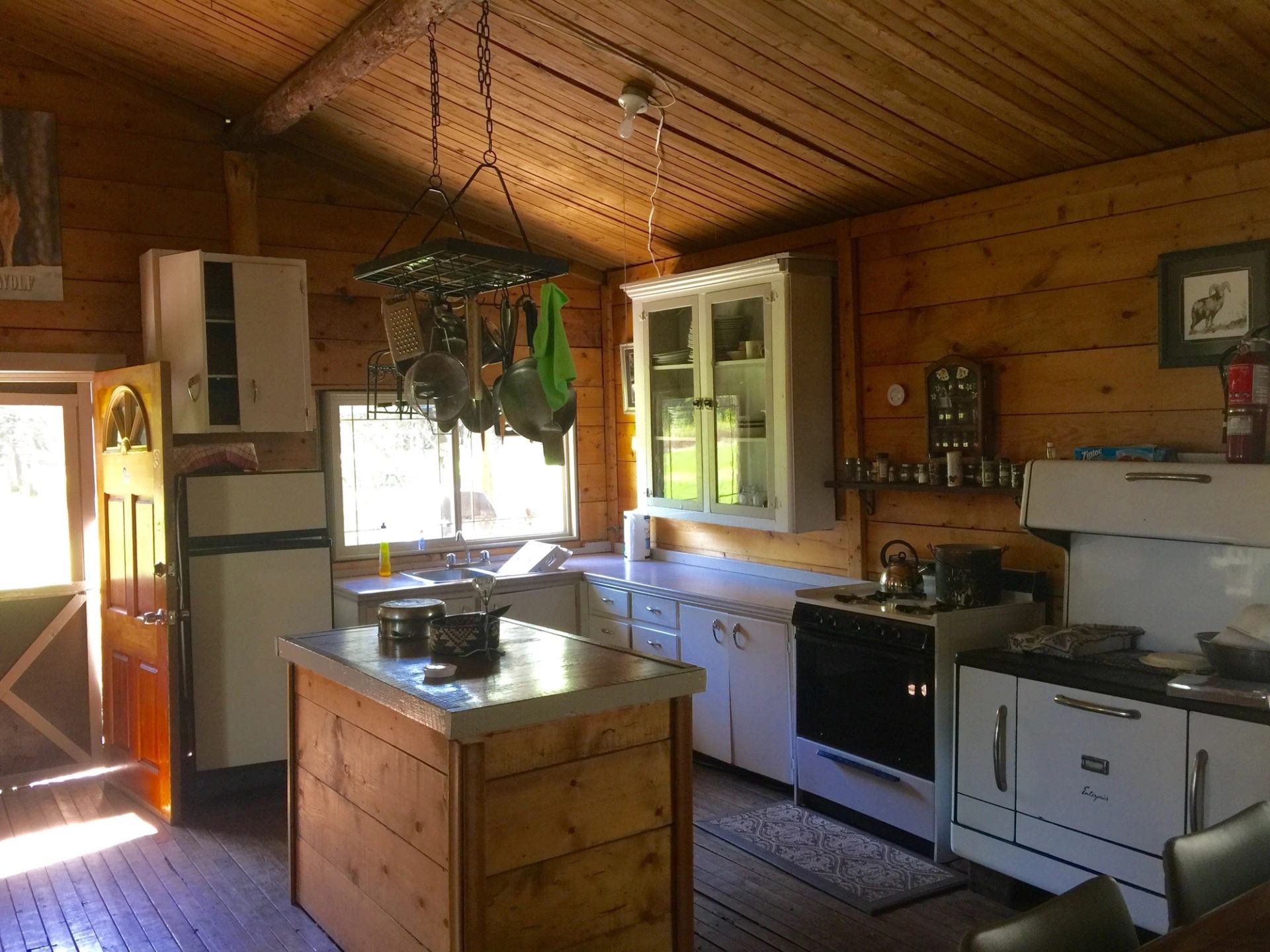 A rustic kitchen with wooden walls, a stove, fridge, hanging pots, and a wooden floor.