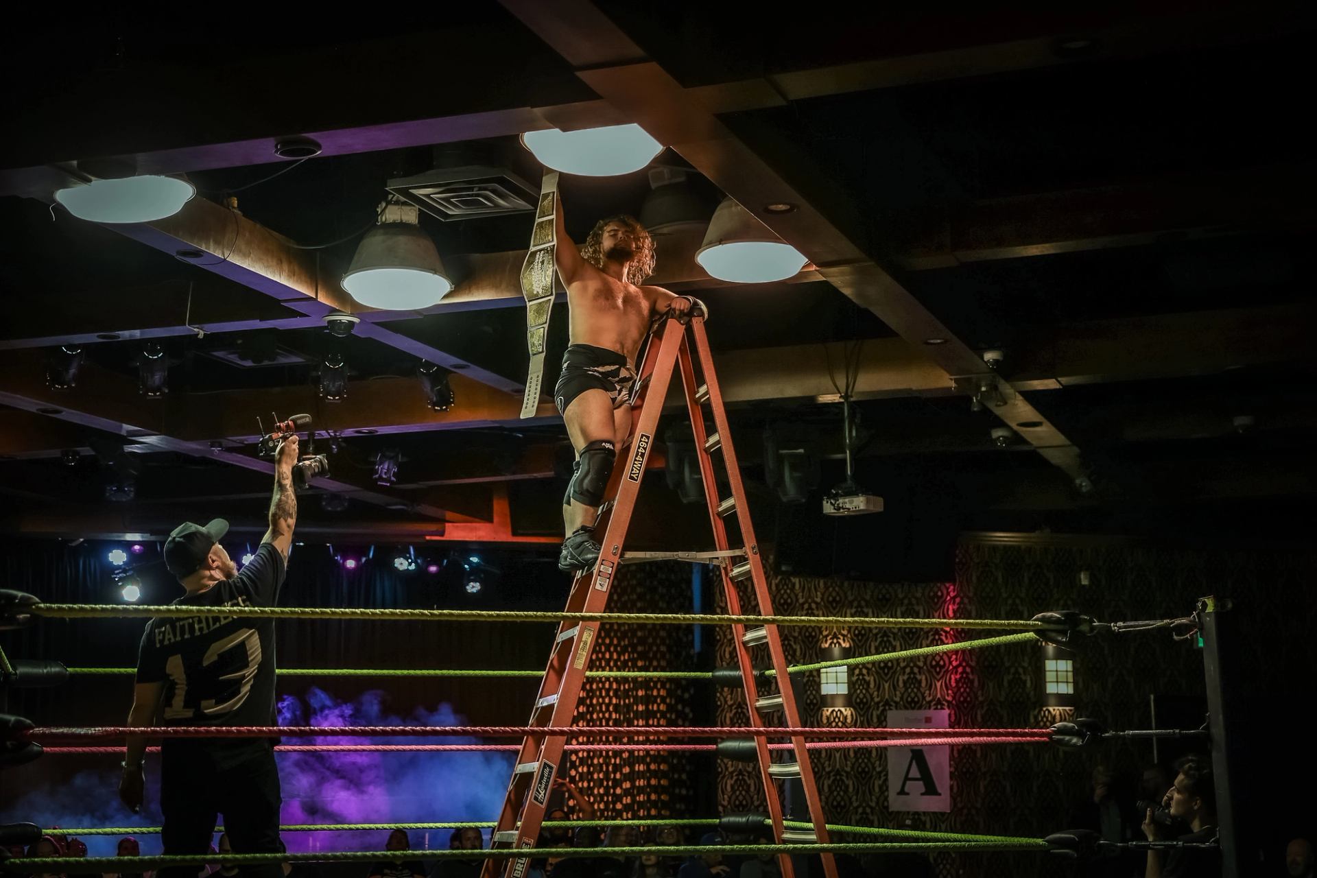 Wrestler stands on a ladder in the ring holding a championship belt.