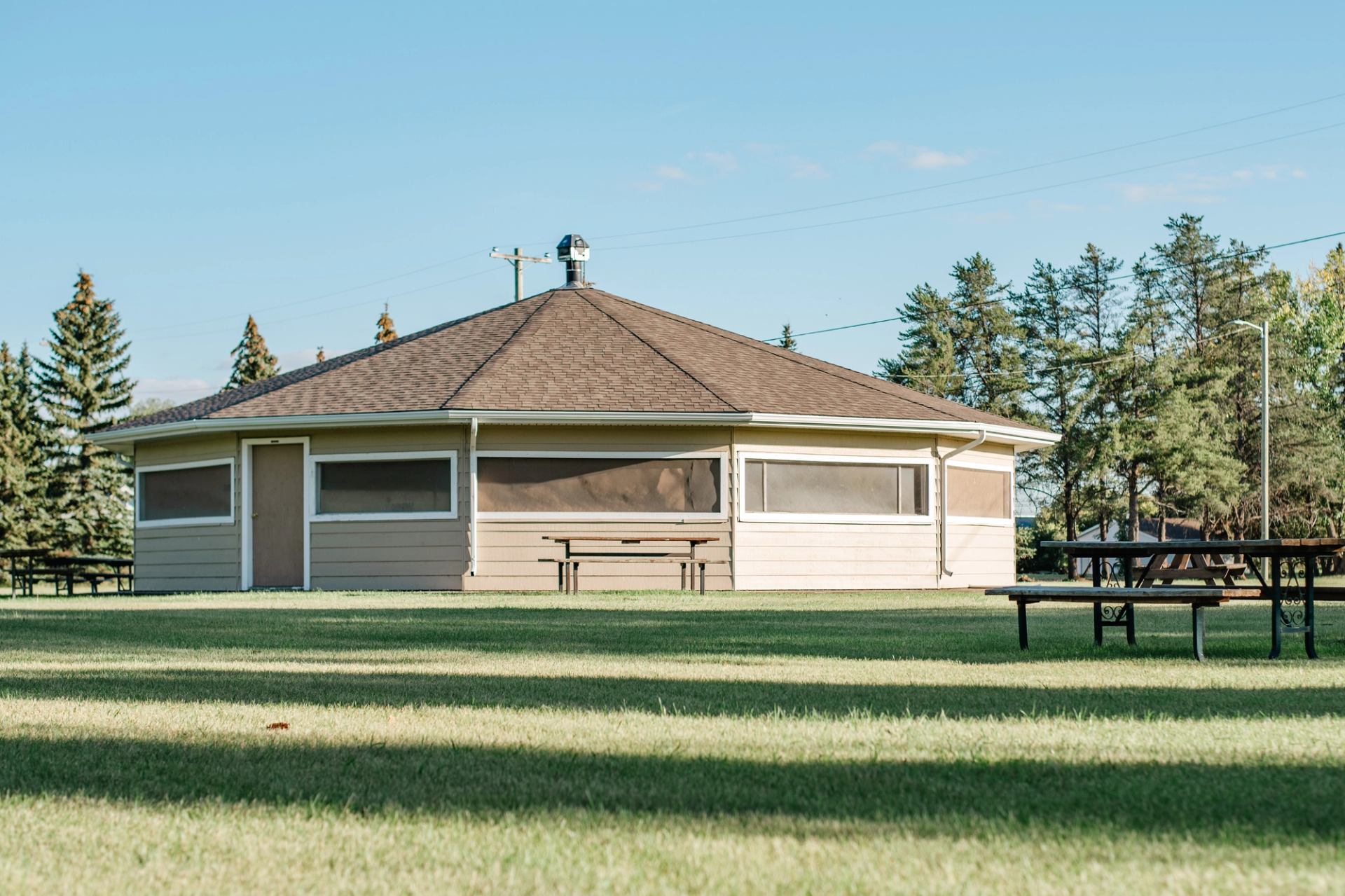 Octagonal park building with brown roof, surrounded by grass, trees and picnic tables.