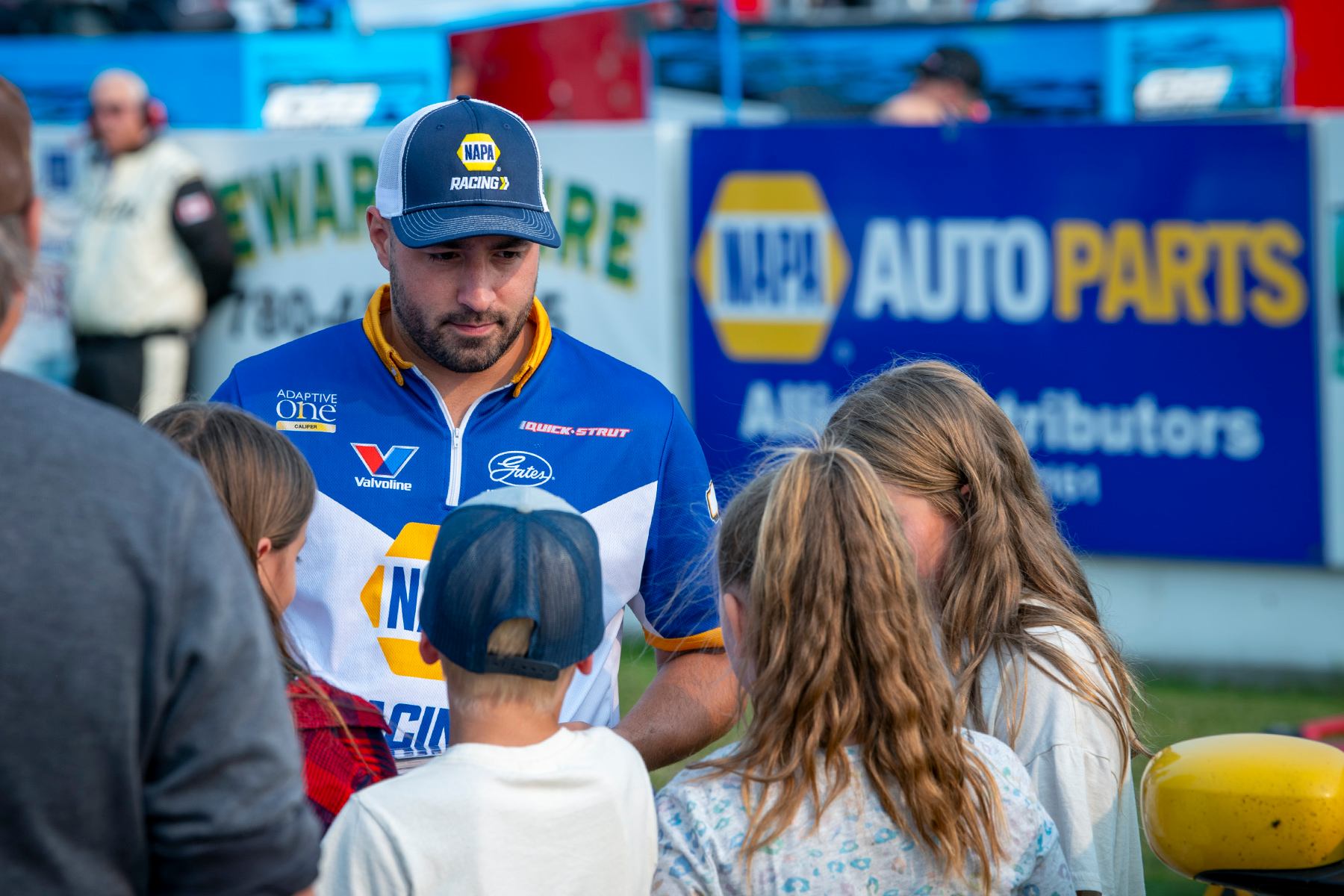 Person in a NAPA-branded shirt talking with a group of children near a NAPA Auto Parts sign.
