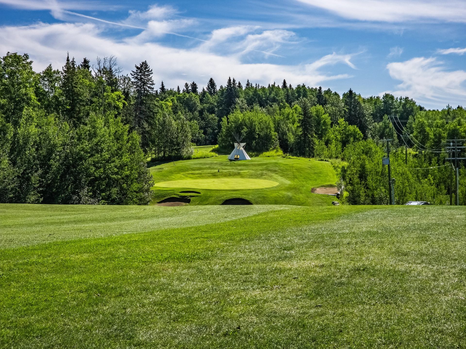 A golf course with a white teepee on a distant green hill, surrounded by forest under a blue sky.