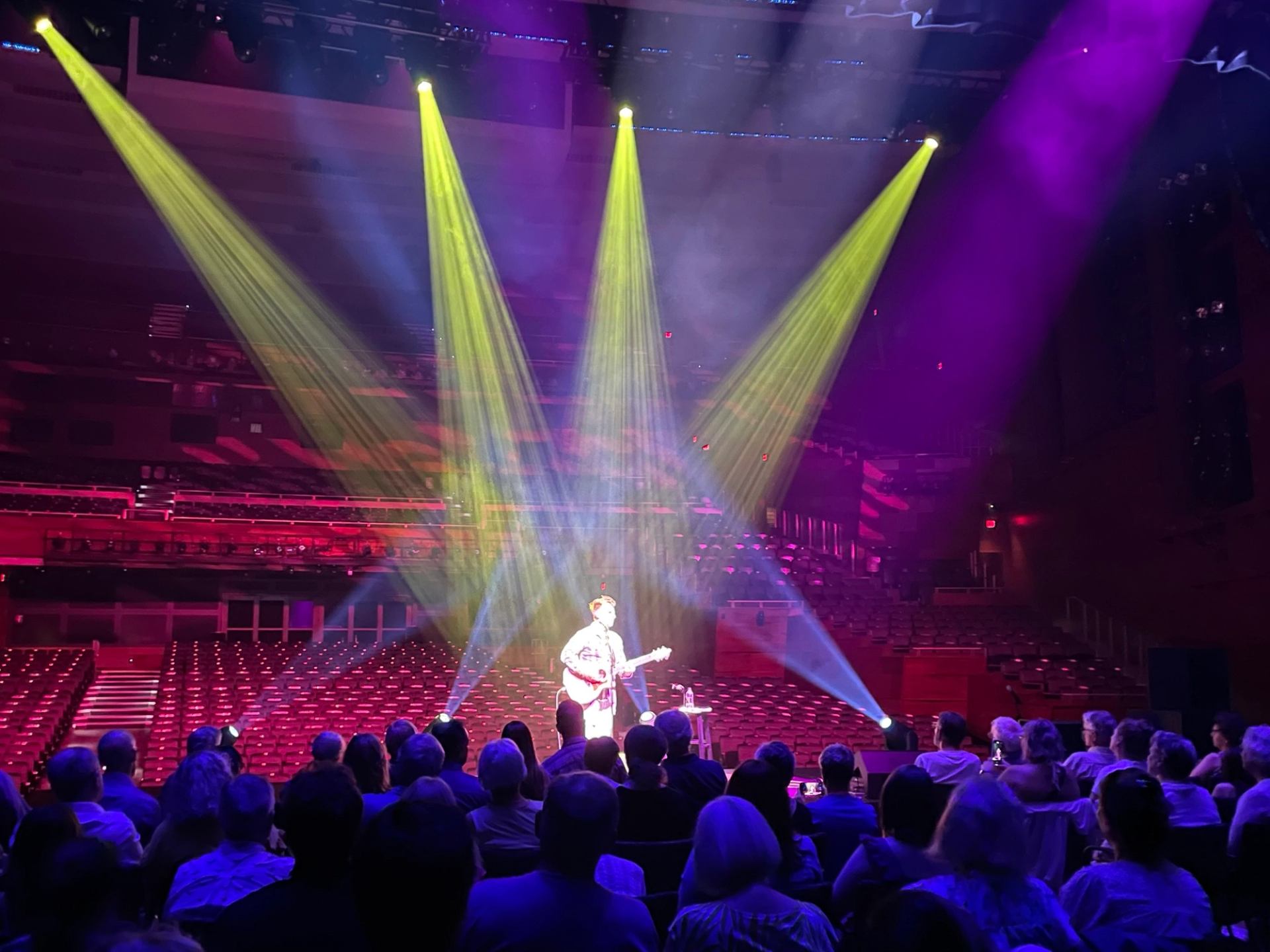 Guitarist performs on stage under colourful spotlights before seated audience in large theatre.