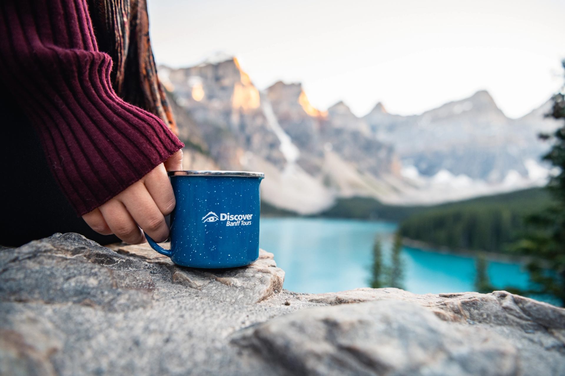 Hand holding blue Discover Banff Tours mug on rock above turquoise mountain lake.