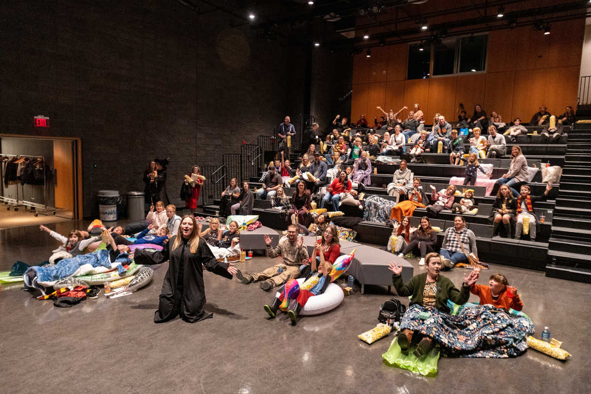 Audience members sit on tiered theatre seating with blankets, cushions and snacks.