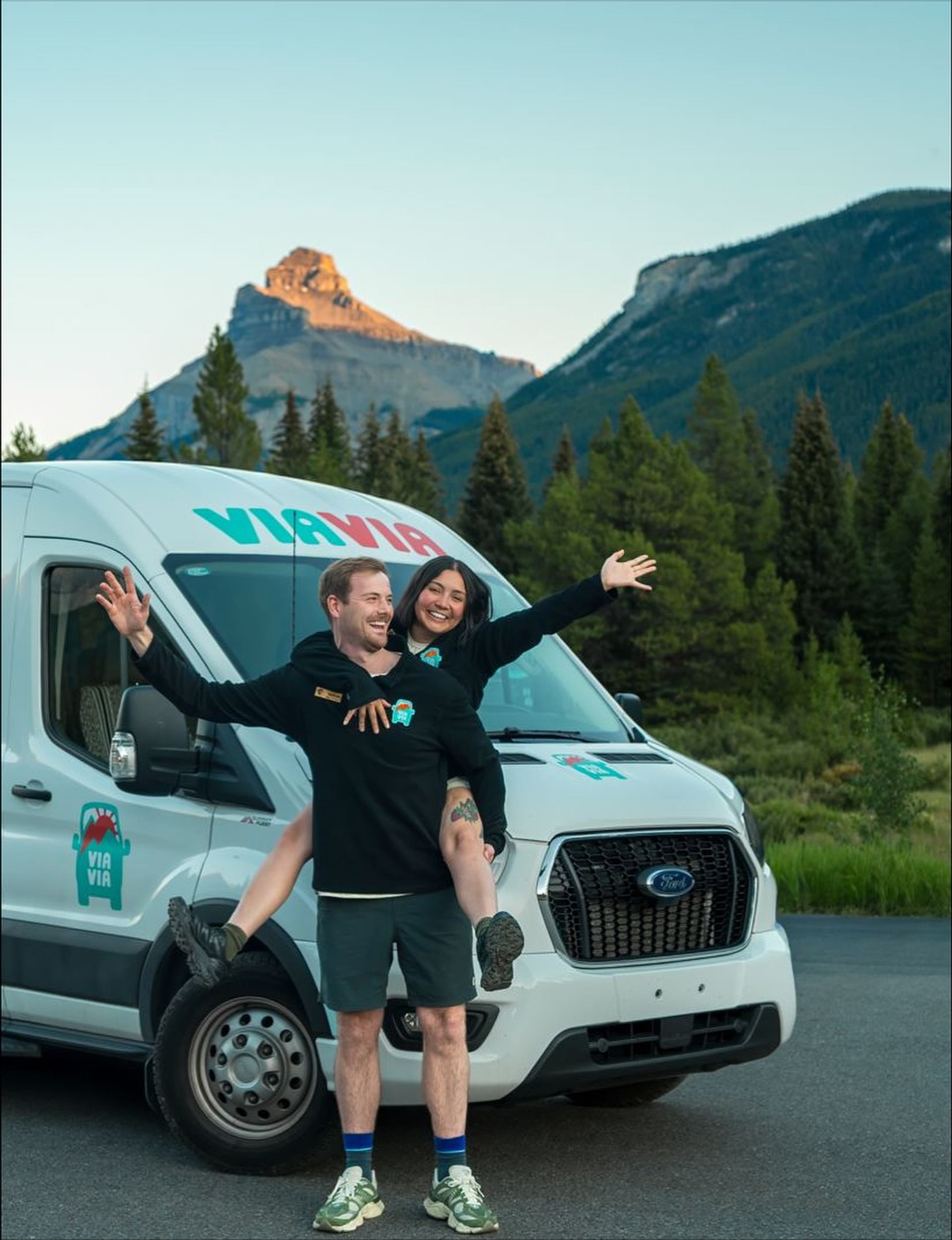 A man gives a piggyback ride to a smiling woman with outstretched arms in front of a white ViaVia van.
