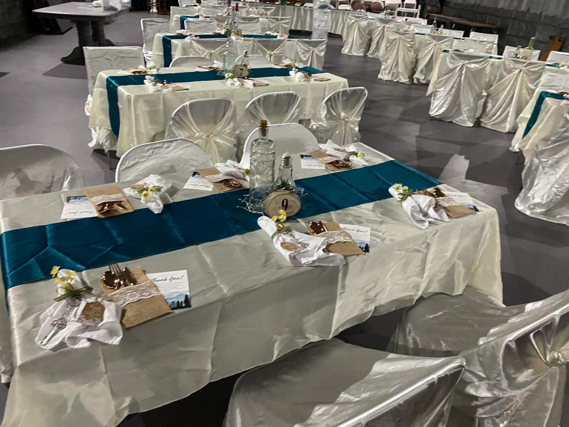 Rows of decorated tables with teal runners and white chair covers arranged for an event.