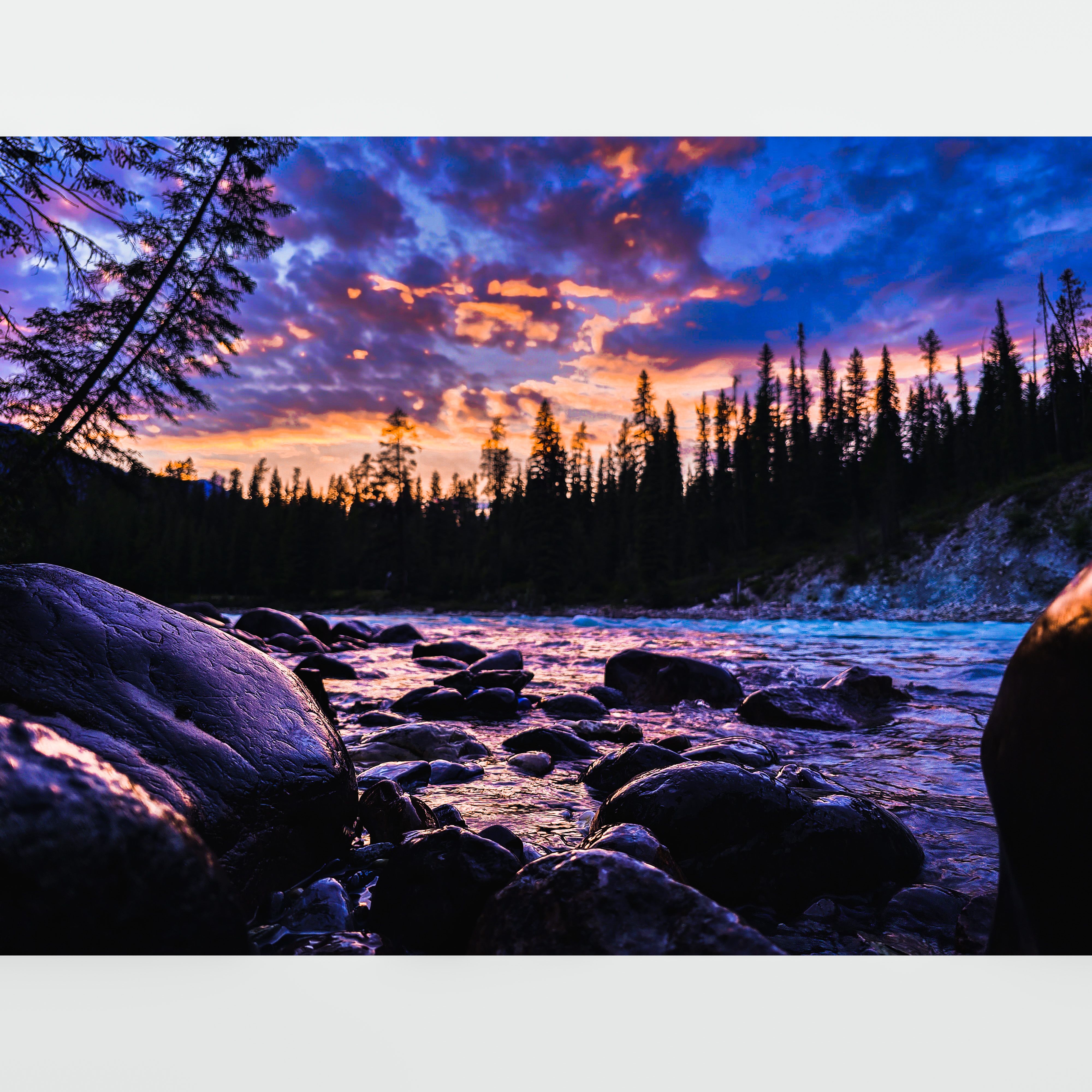 Sunset casts pink and purple hues over a forest river, with smooth stones and calm, reflective water.