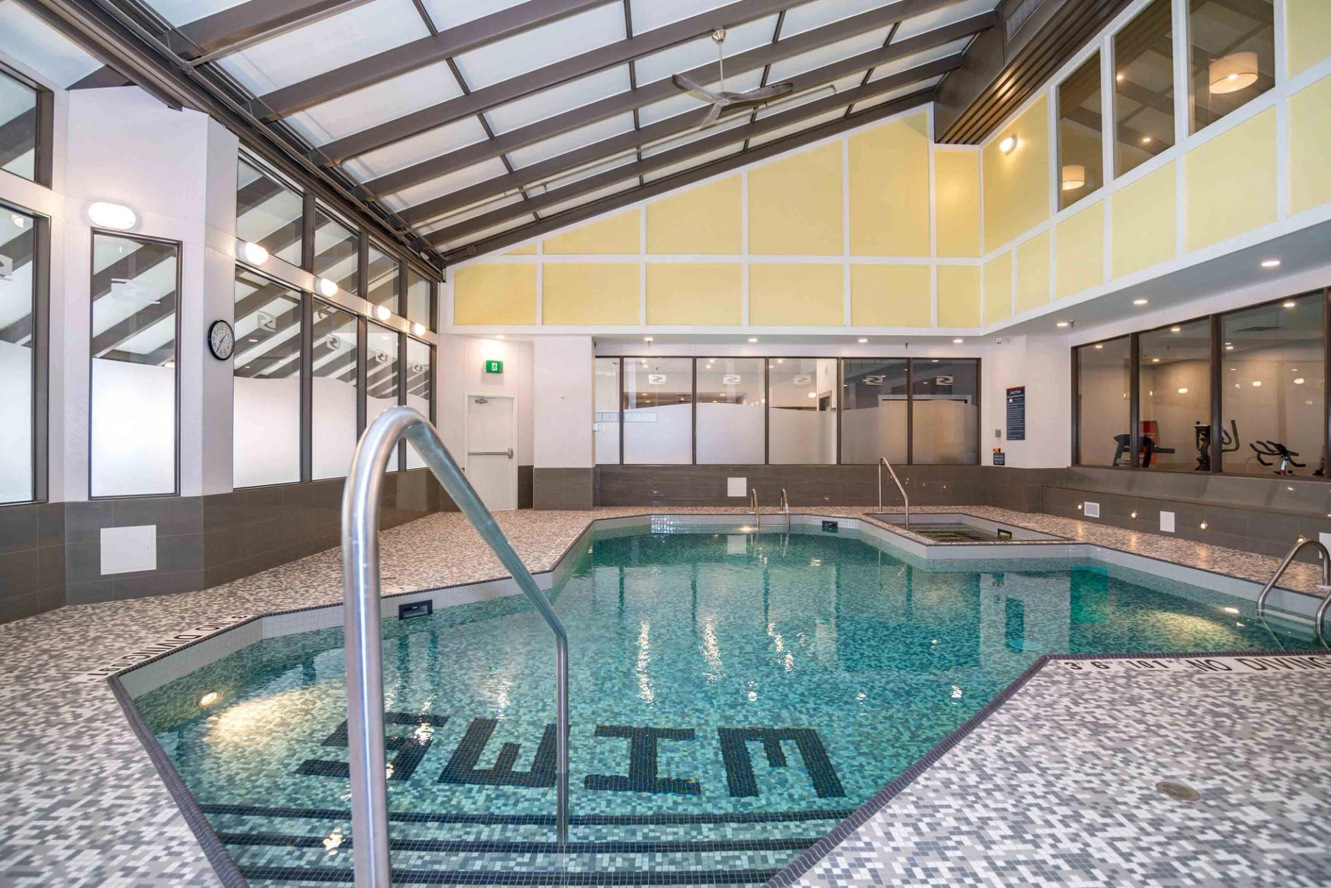 Indoor swimming pool with tiled deck, glass walls, hot tub and fitness room beyond.