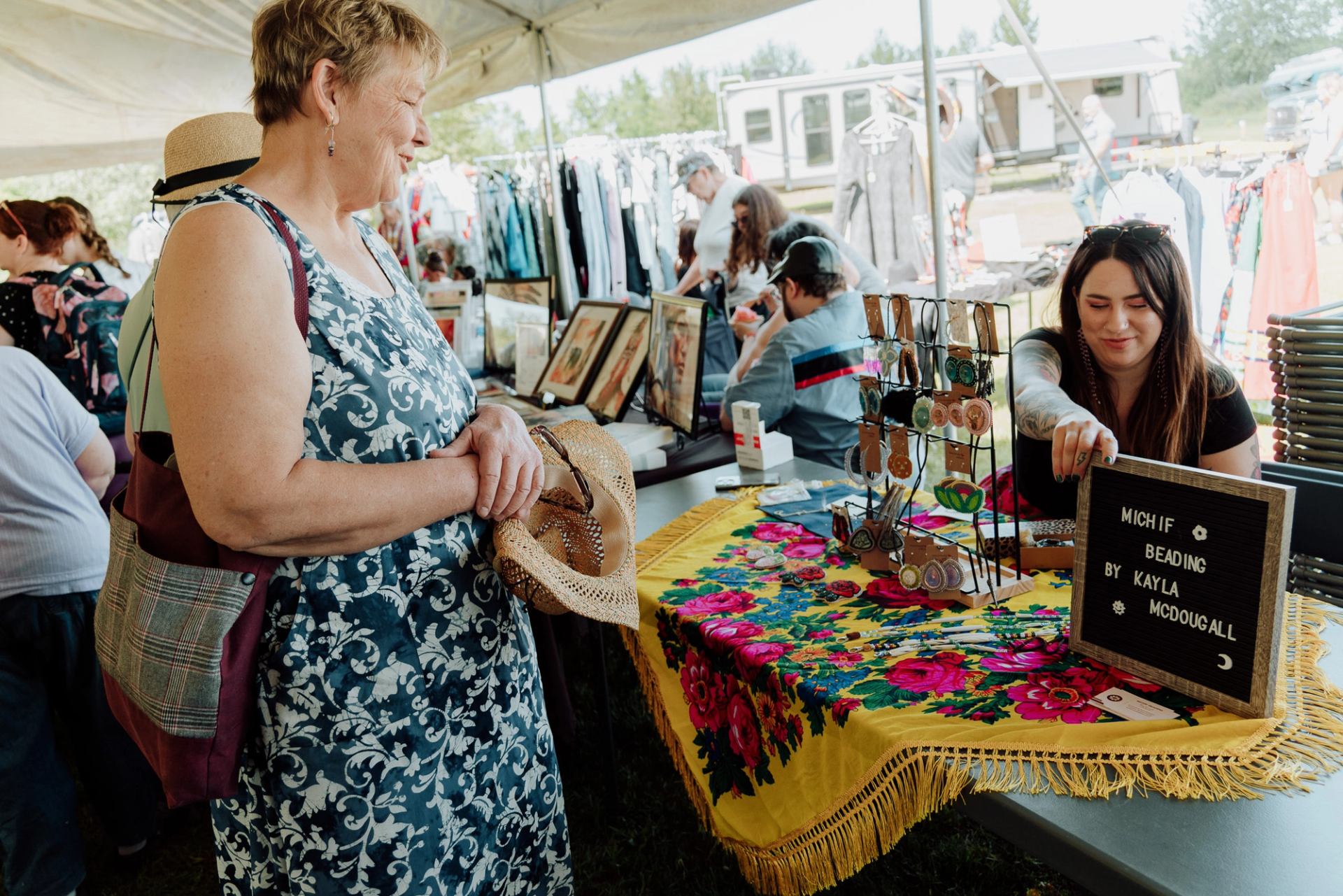 Market vendor arranging beaded jewellery display as shopper browses colourful table under tent.