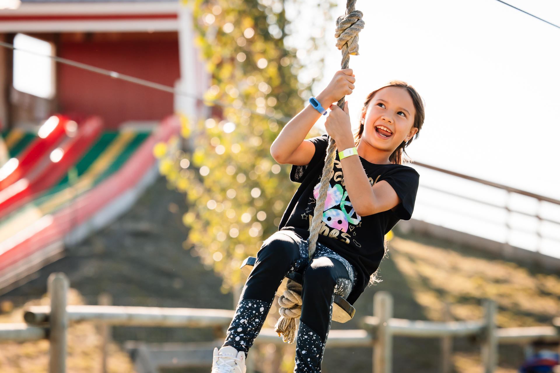 Child on rope swing at outdoor park, with colourful slides in background.