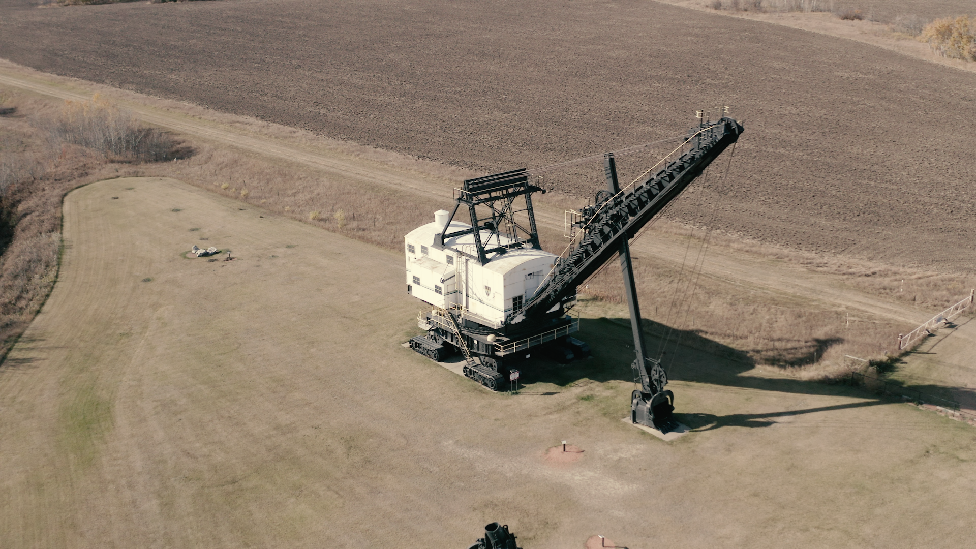 Large dragline excavator sits on open grassy field beside ploughed farmland and dirt road.