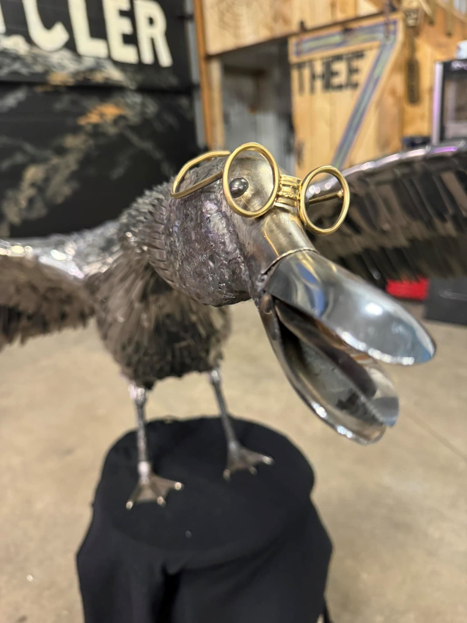 Metal bird sculpture with round glasses and open beak on black pedestal.