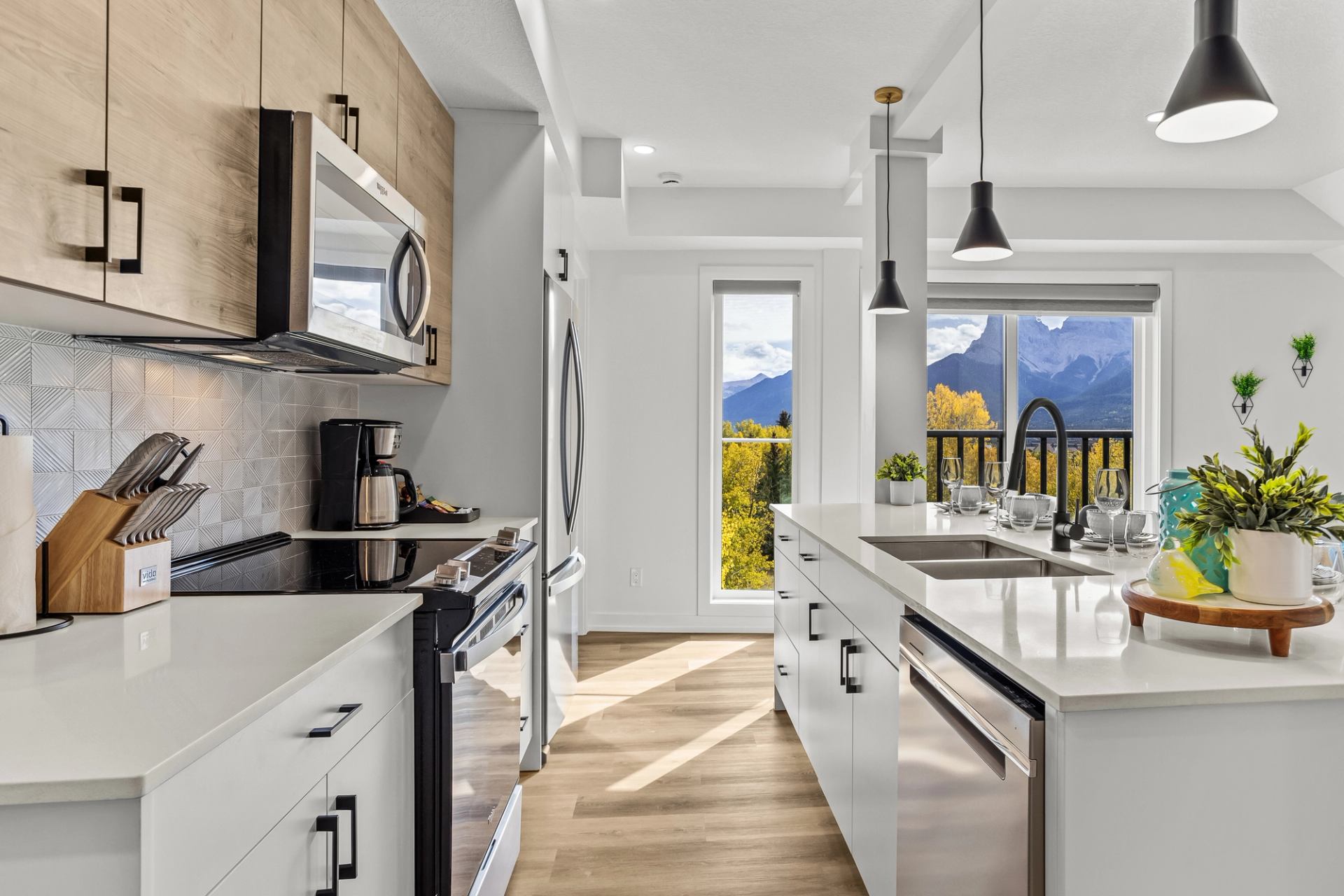 Modern white kitchen with island, stainless appliances and mountain view through large windows.