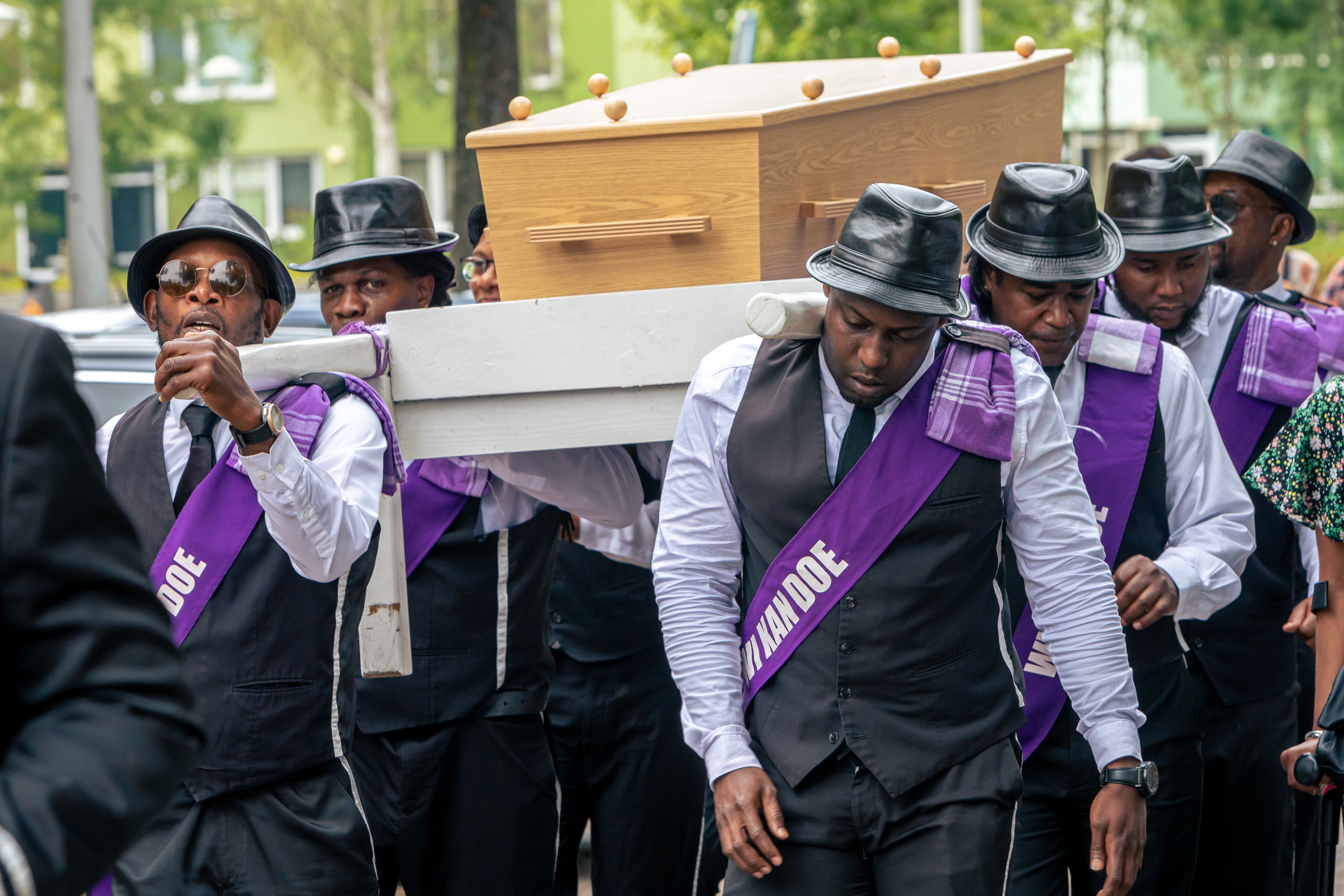 Surinaamse dragers in wit overhemd, zwart gilet, zwarte hoed en paarse sjerp dragen samen een lichte houten uitvaartkist op een witte draagbaar.