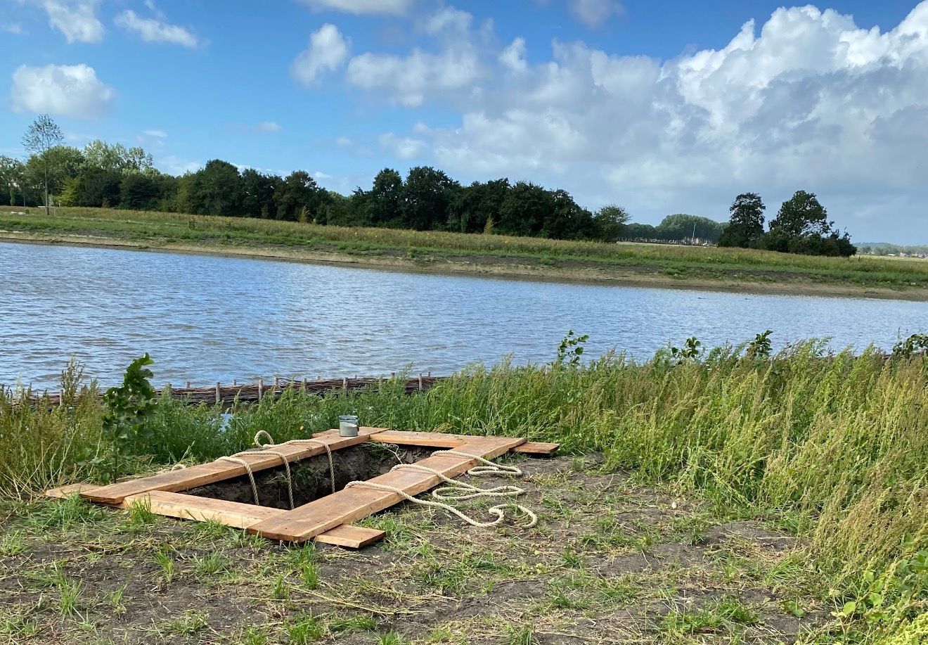 Een vers gegraven natuurgraf aan de oever van een breed water, met een raamwerk van houten planken en dikke touwen over de opening. Eromheen hoog gras en wilde planten, aan de overkant een rij bomen onder een blauwe lucht met witte wolken.