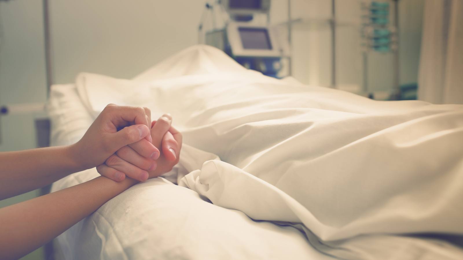 Twee handen die elkaar vasthouden op de rand van een ziekenhuisbed