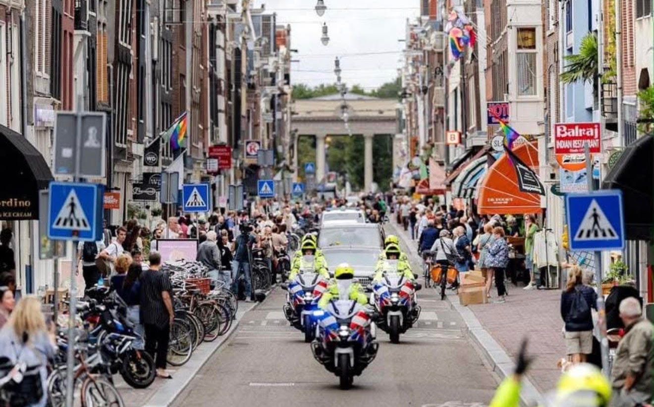 De rouwstoet in de Haarlemmerstraat in Amsterdam: vier politiemotoren rijden voor de rouwauto uit, op de stoepen staan mensen te kijken, met in de verte de zuilen van de Haarlemmerpoort.