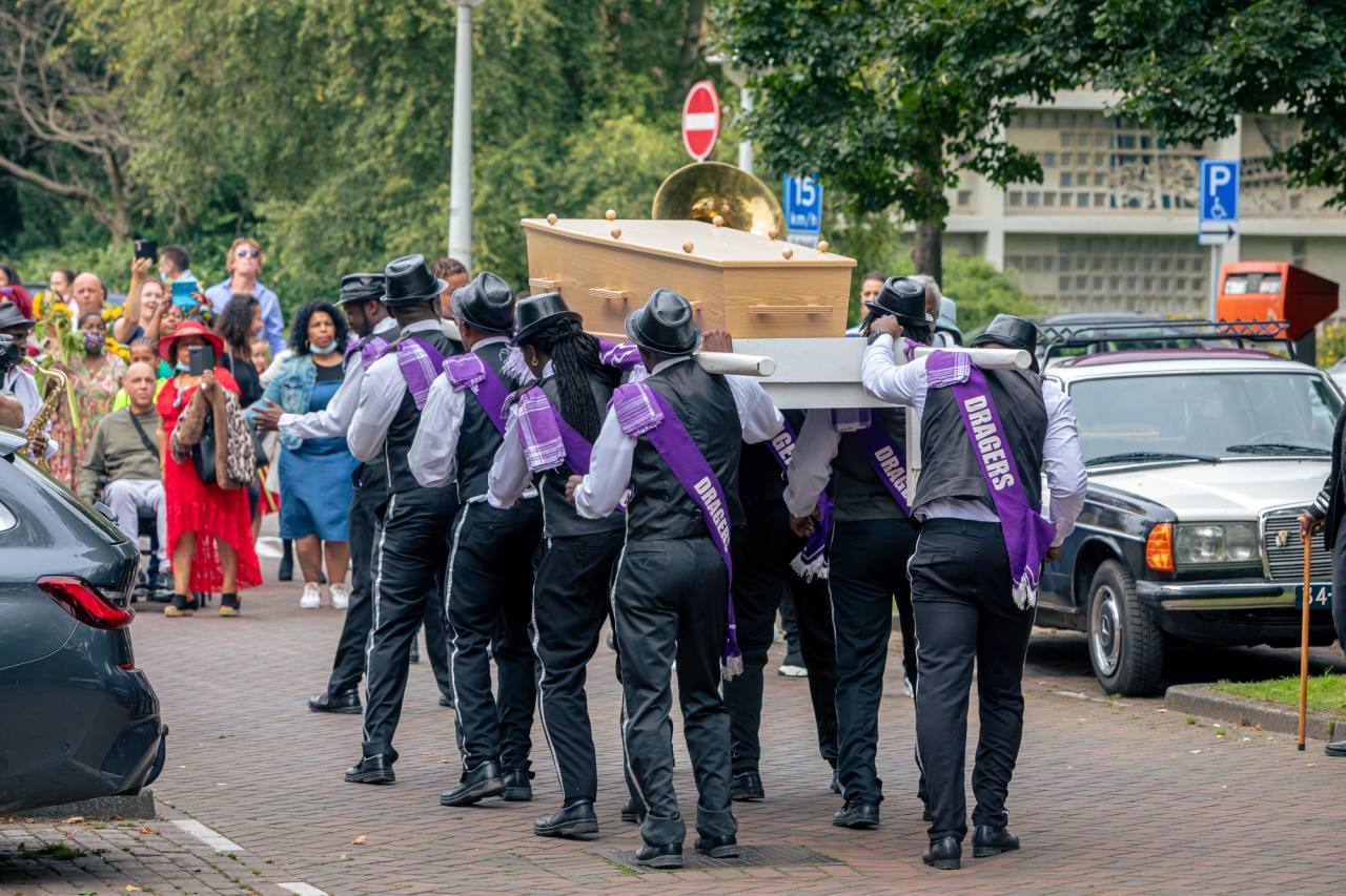 Dragers met paarse sjerpen lopen van achteren gezien met de uitvaartkist over straat; boven de kist steekt de koperen klok van een bazuin uit en langs de route kijken familie en vrienden toe.