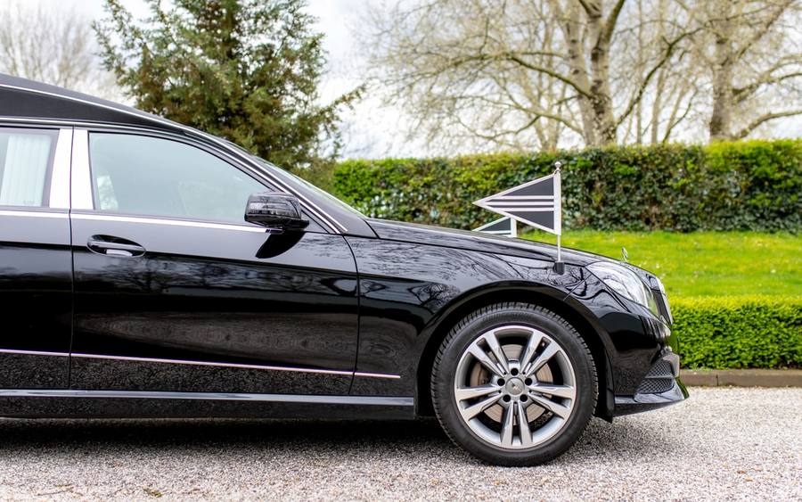 Een zwarte rouwauto met een wimpel op de motorkap, langs een geschoren heg