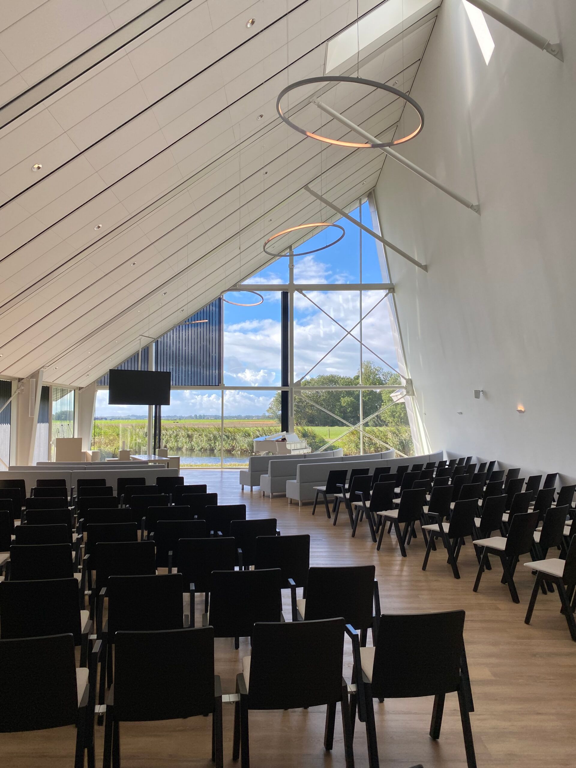 De aula van Crematorium Alkmaar, met een hoog schuin dak en een glaswand met uitzicht op de weilanden