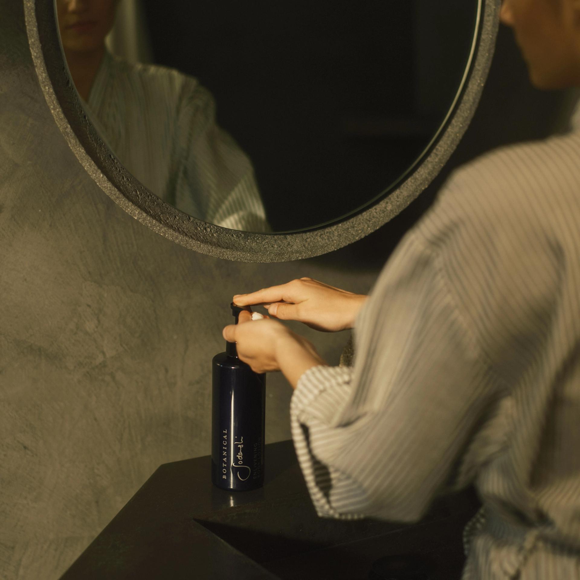 Woman in robe using Sodashi Body Lotion dispenser on bathroom vanity