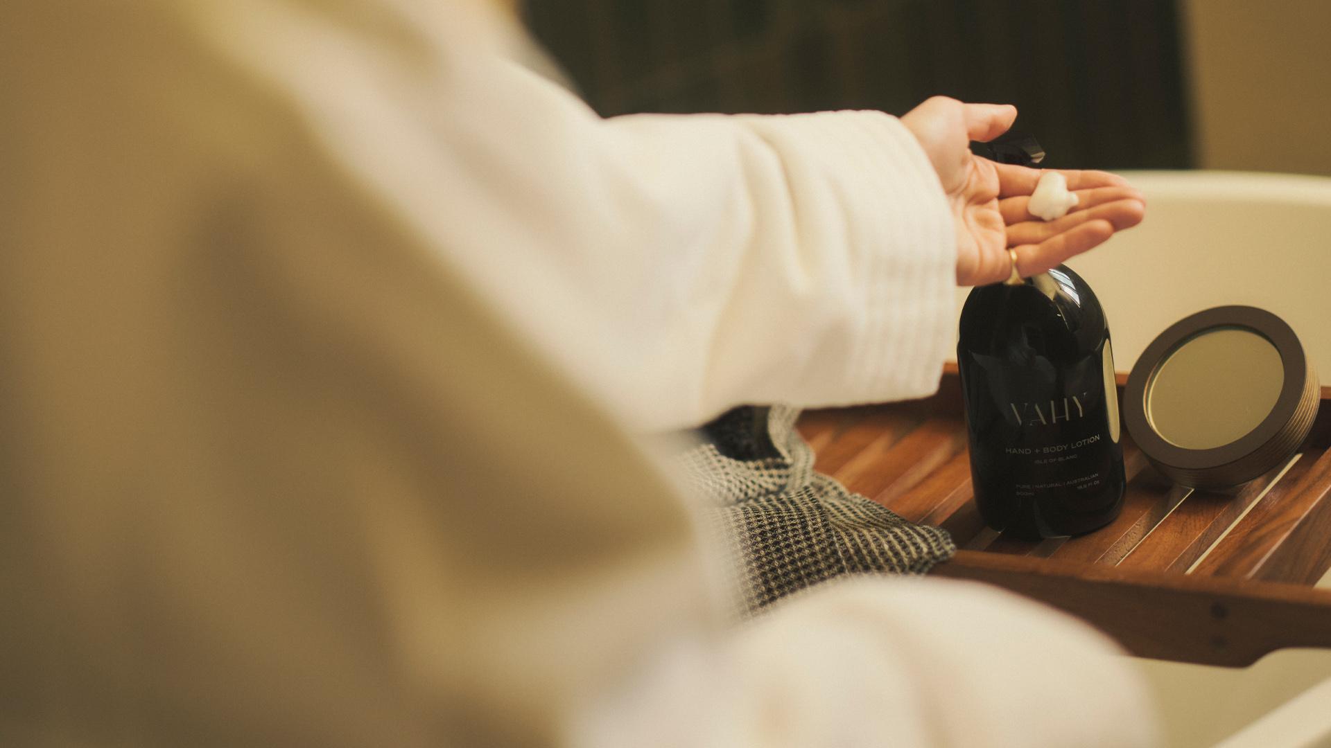 Woman in robe using Vahy Isle of Blanc Body Lotion dispenser on bathtub tray