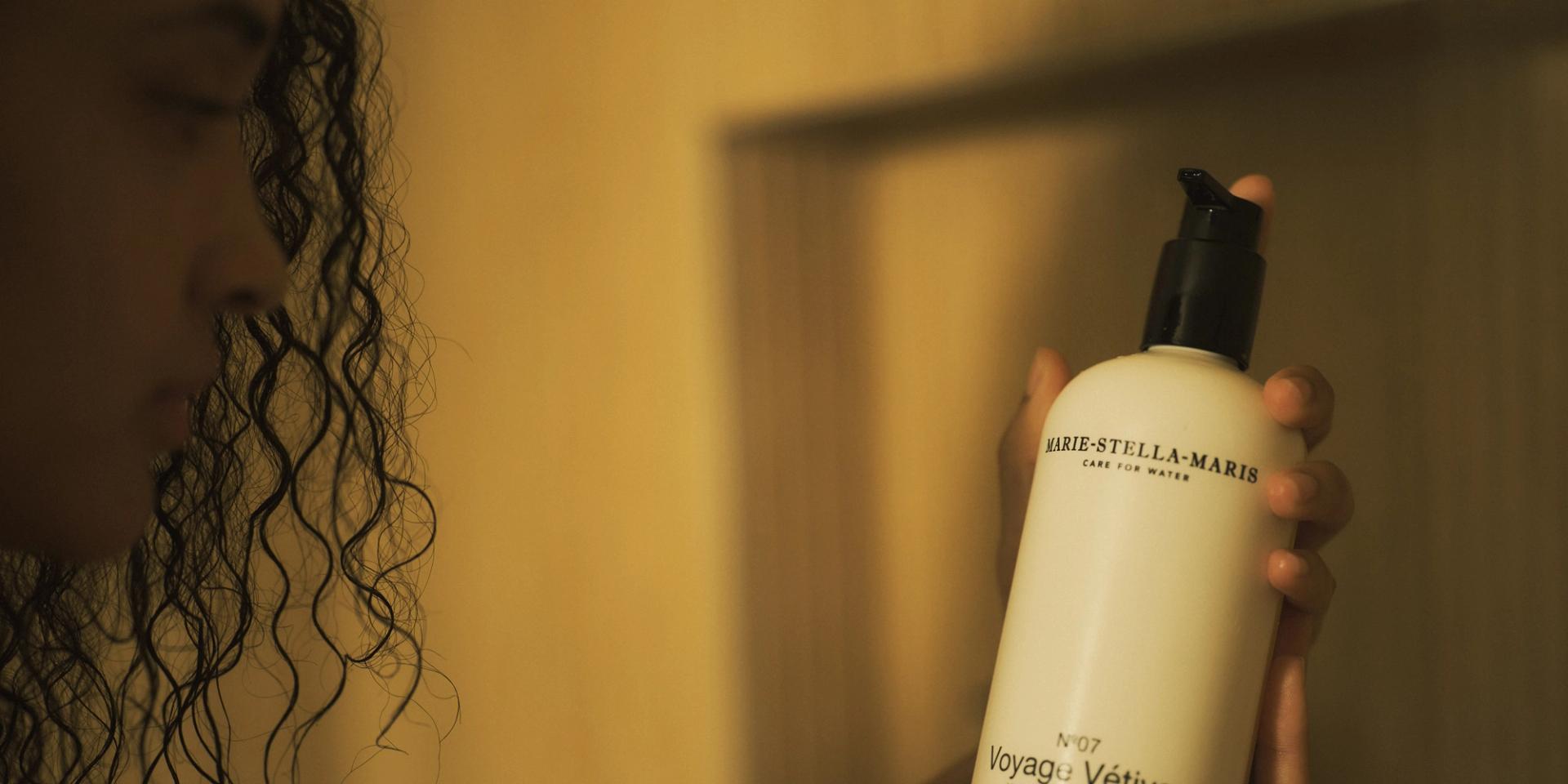 Woman in shower holding Marie Stella Maris Voyage Vetiver Shampoo dispenser in hands