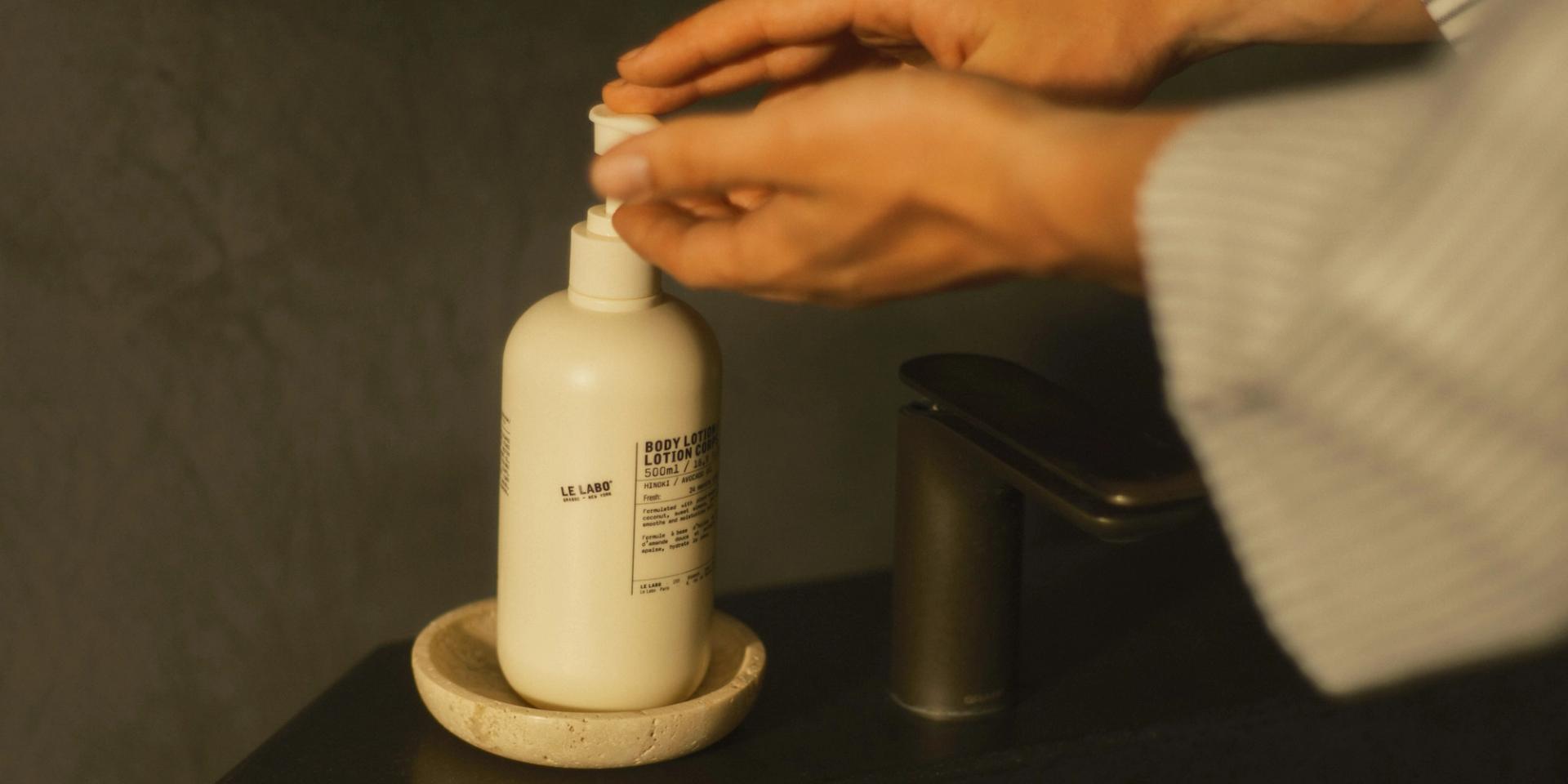 Woman in bathroom using Le Labo Hinoki Body Lotion dispenser on bathroom vanity