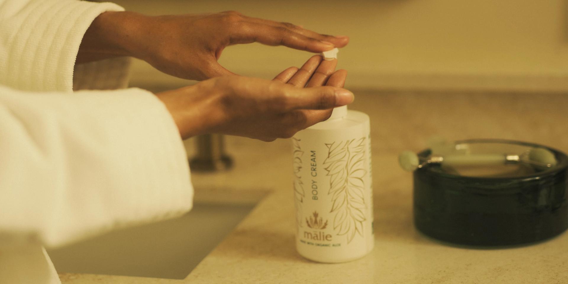 Woman in robe using Malie Koke'e Body Cream dispenser on bathroom vanity