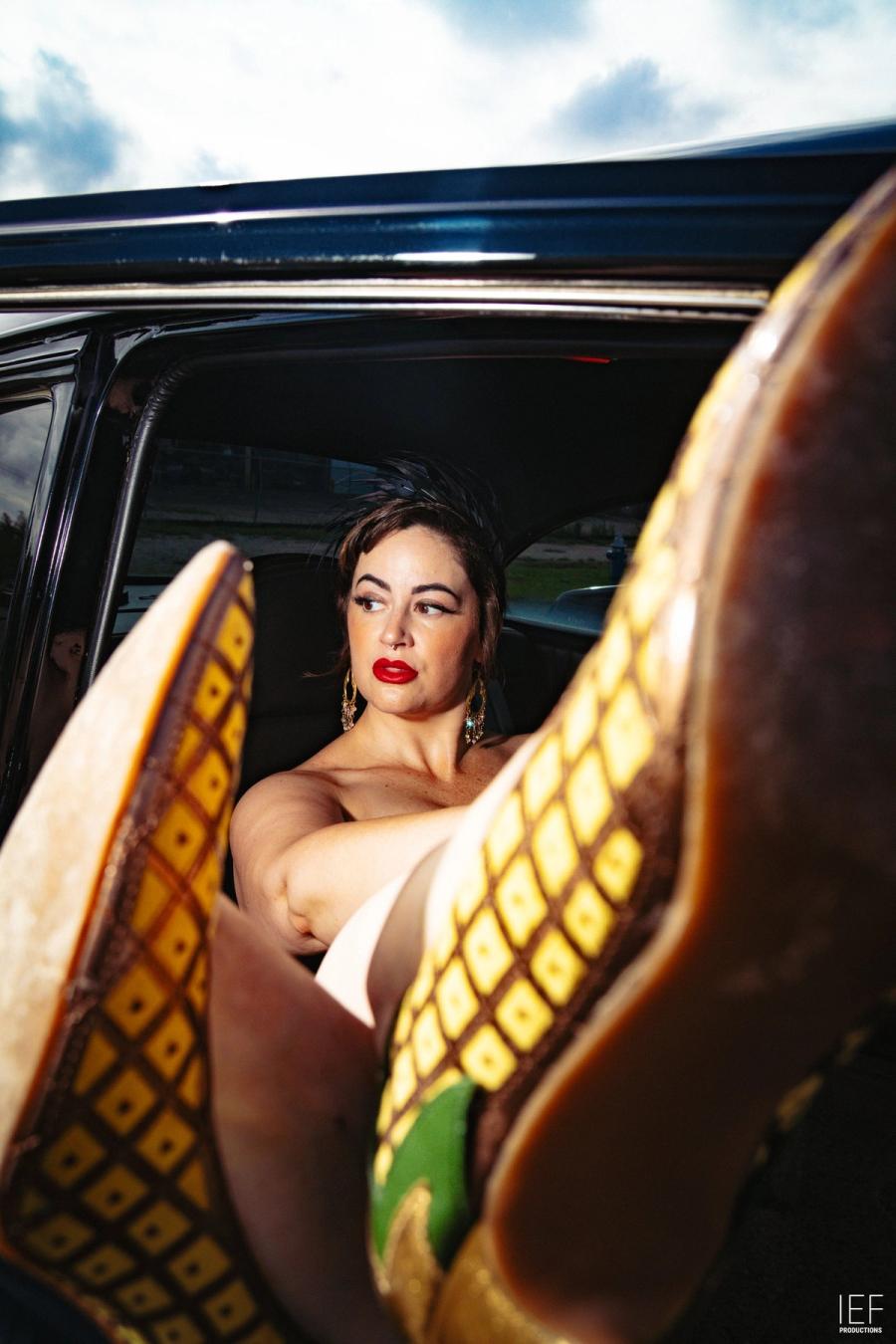A woman with red lips looks out from the back seat of a car, the gold-patterned soles of her boots raised toward the camera
