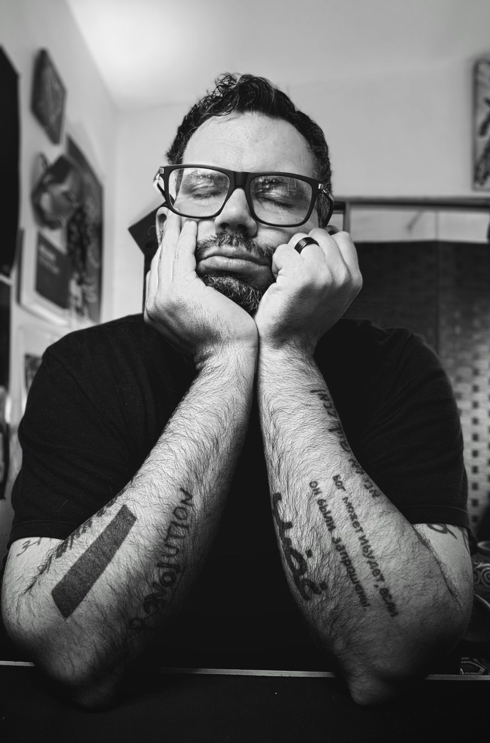 Ian Findley in black-framed glasses and a black T-shirt, chin resting on his hands, tattooed forearms, in black and white