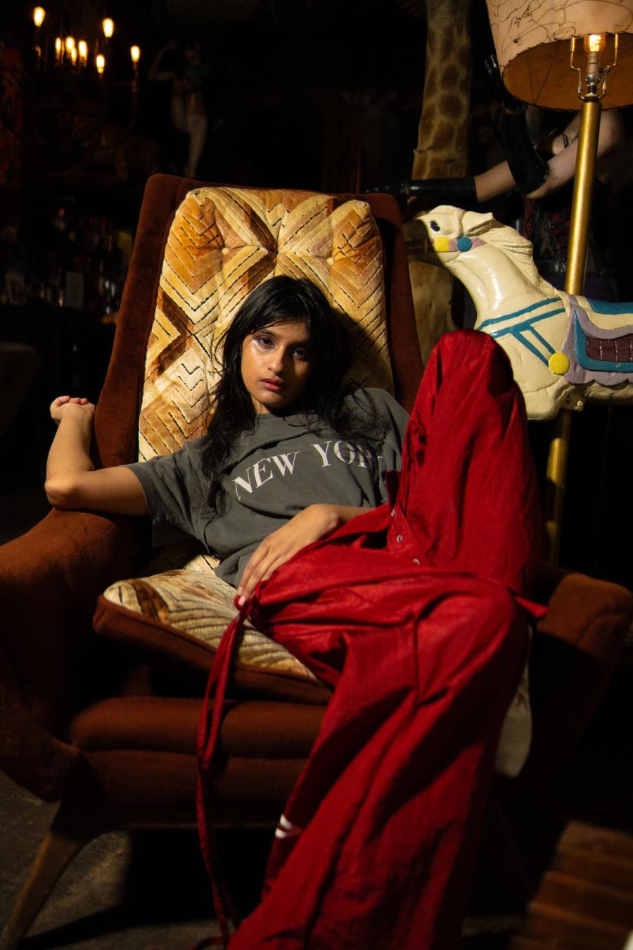 A woman in a grey New York T-shirt lounging in a patterned armchair under a red robe, a carousel horse and a lamp behind her