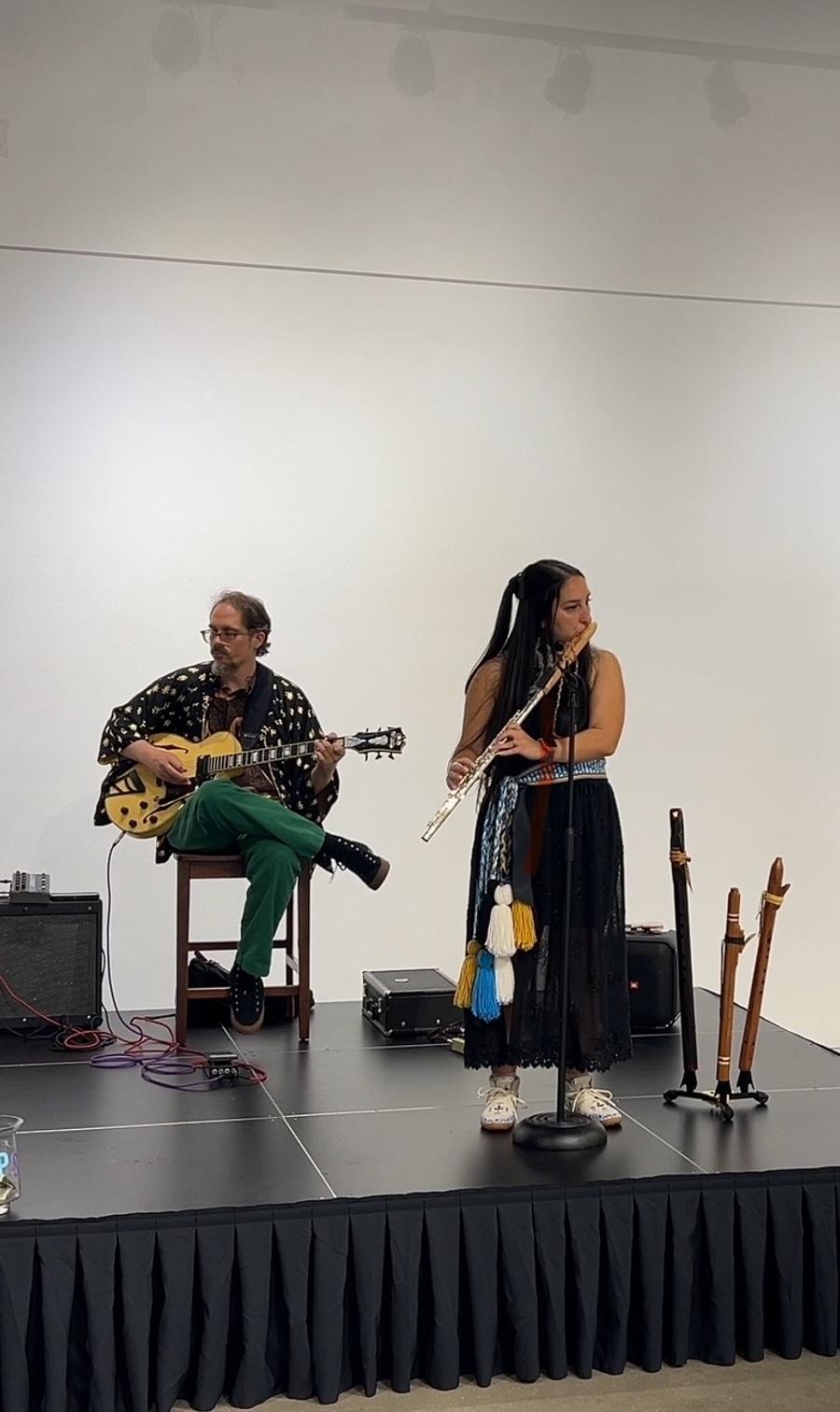 Aryn Ward playing flute on a small stage beside a seated guitarist, three cedar flutes on stands