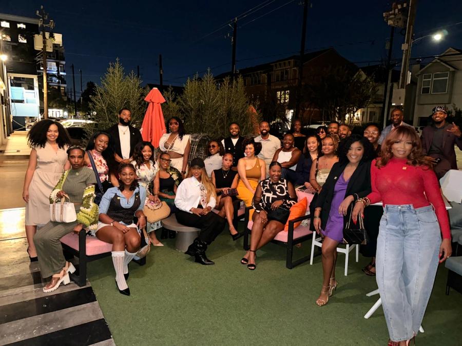 A large group of Black creatives gathered on a lawn at an evening event, city buildings and string lights behind them
