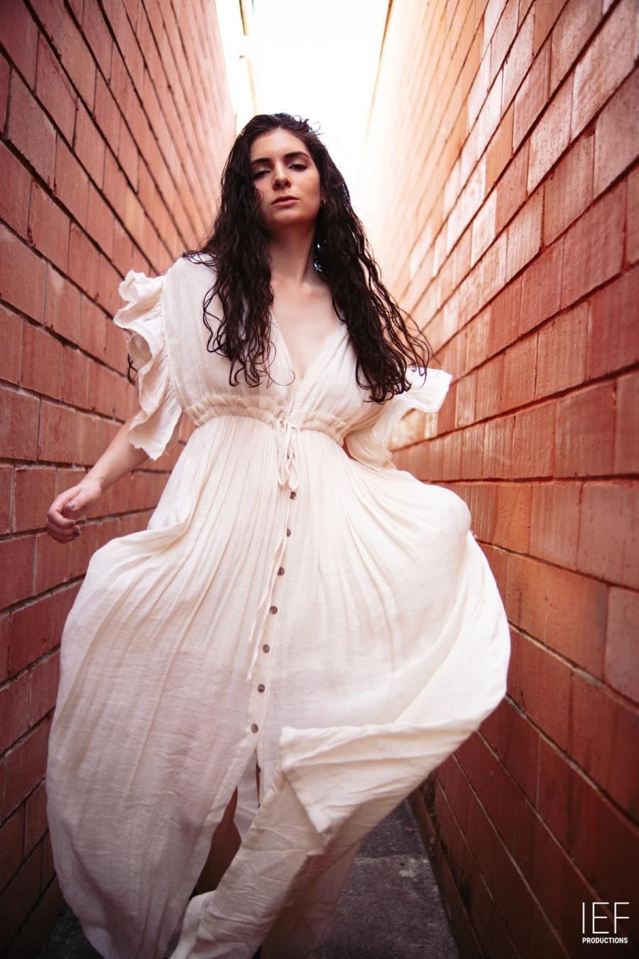 A woman with long dark curls in a flowing white buttoned dress walking between two red-brick walls