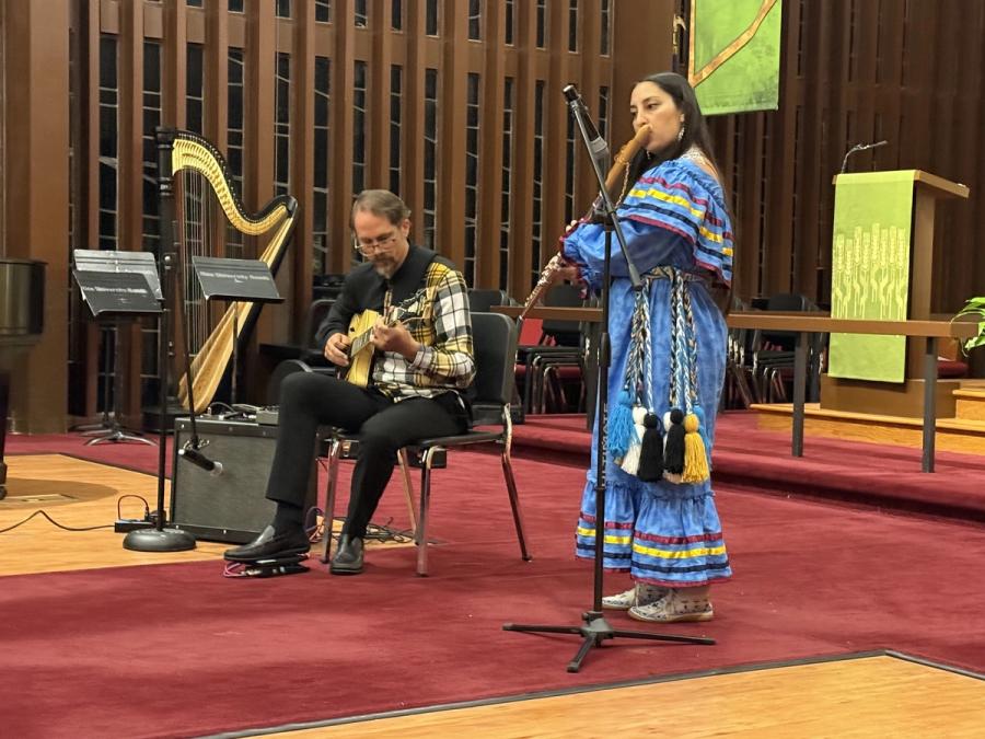 Aryn Ward in a blue ribbon dress playing a cedar flute in a church sanctuary, a guitarist and a harp behind her