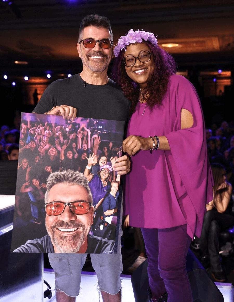 Simon Cowell and Beverly Springer on the America's Got Talent stage holding Johnny Green's painting of Simon taking a selfie with a crowd