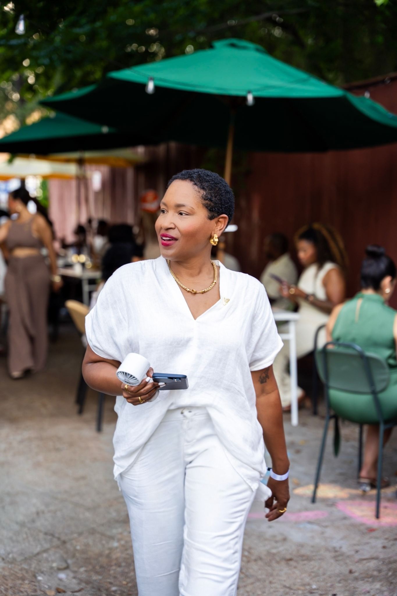 Denise Upshaw in a white linen top and gold necklace, walking across an outdoor patio under green umbrellas