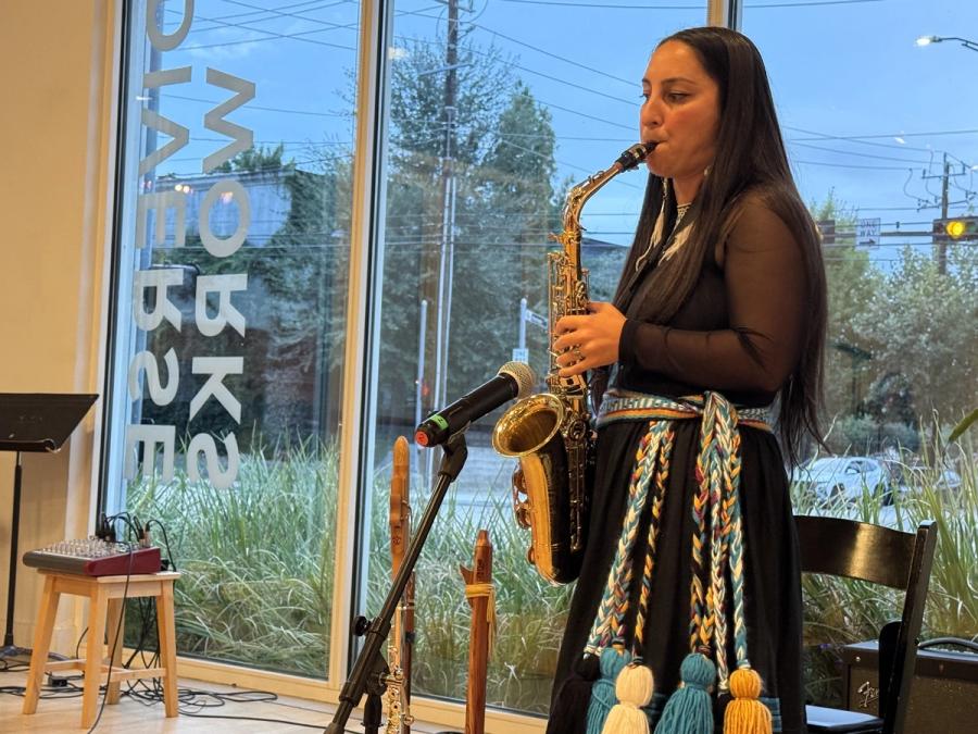 Aryn Ward playing alto saxophone at a microphone, wooden flutes standing beside her, a tall window behind