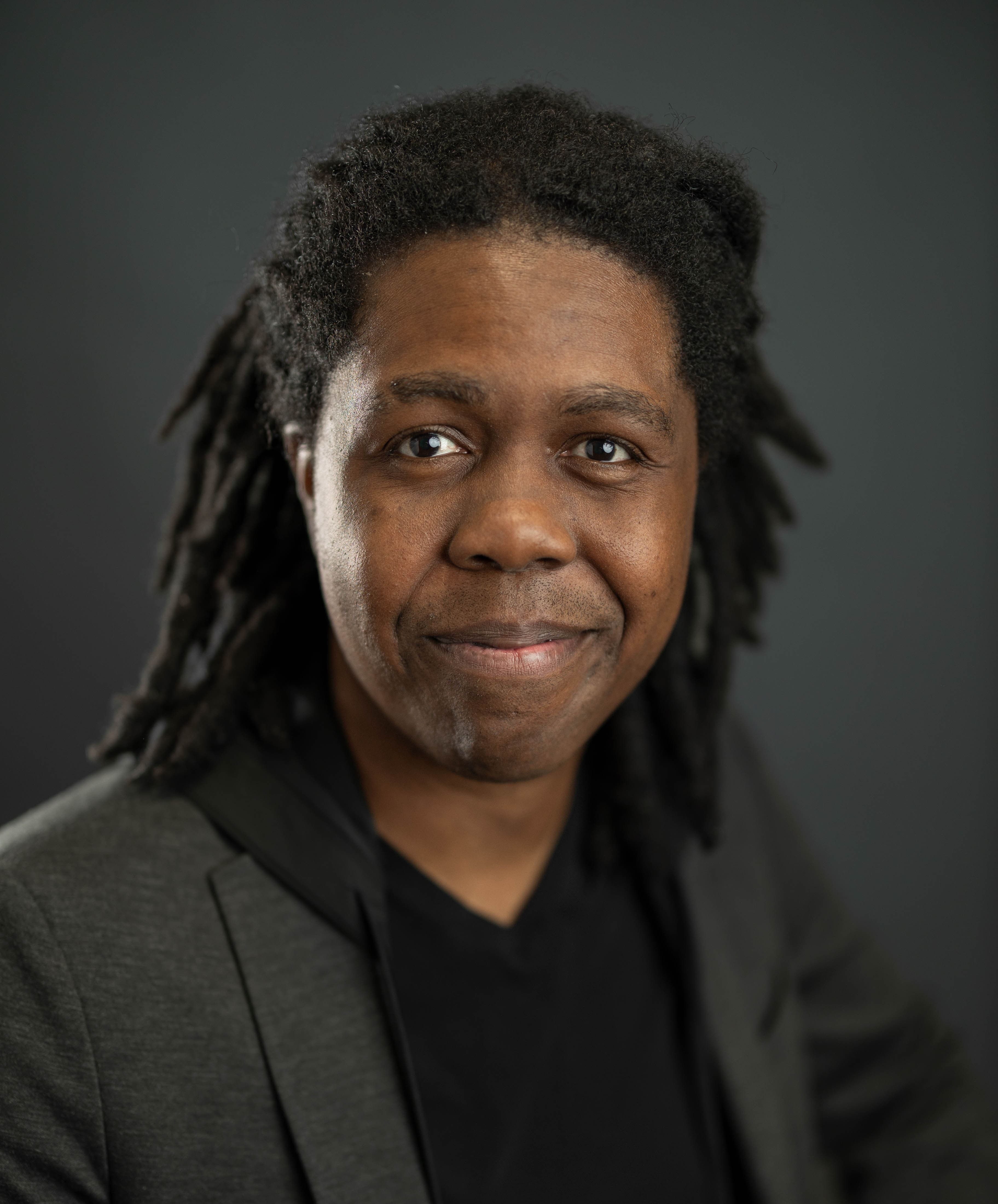 Gary Bedell smiling in a dark jacket and black T-shirt, shoulder-length locs, against a dark grey studio backdrop