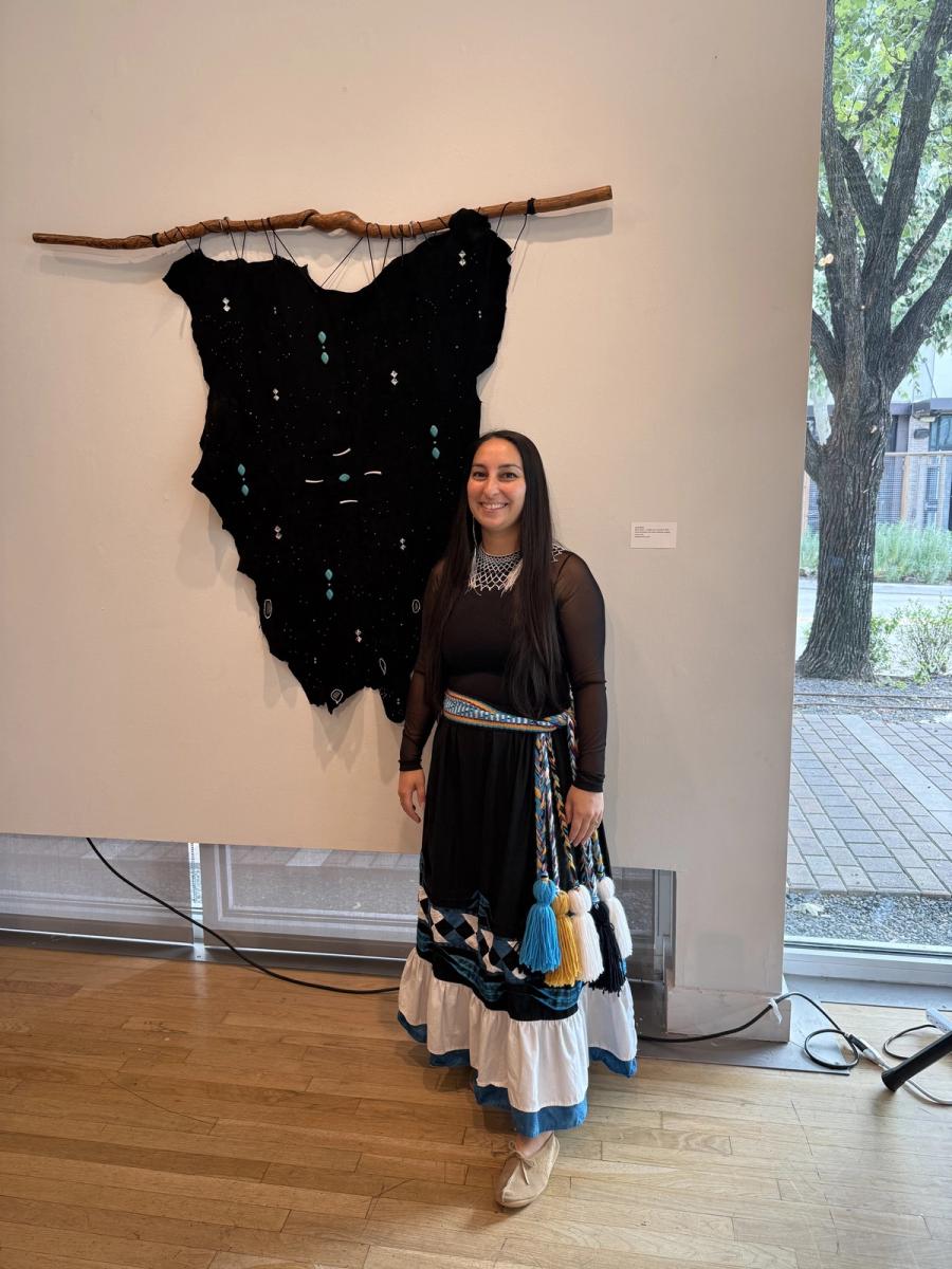 Aryn Ward standing in a gallery beside Yuchi Galaxy, a black deer hide beaded with constellations and hung from a wooden walking stick