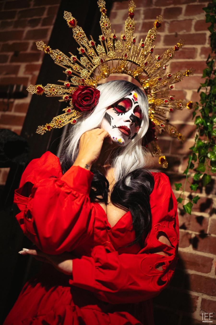 A woman with white hair and sugar-skull face paint in a red dress and a gold sunburst crown with red roses, against a brick wall