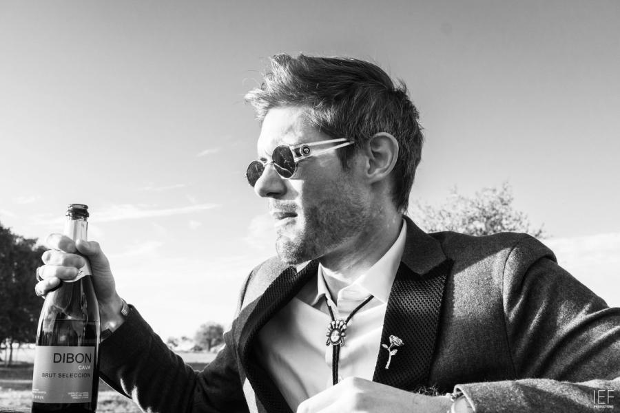 Black-and-white photo of a man in sunglasses and a suit jacket with a bolo tie holding up a bottle of sparkling wine under an open sky
