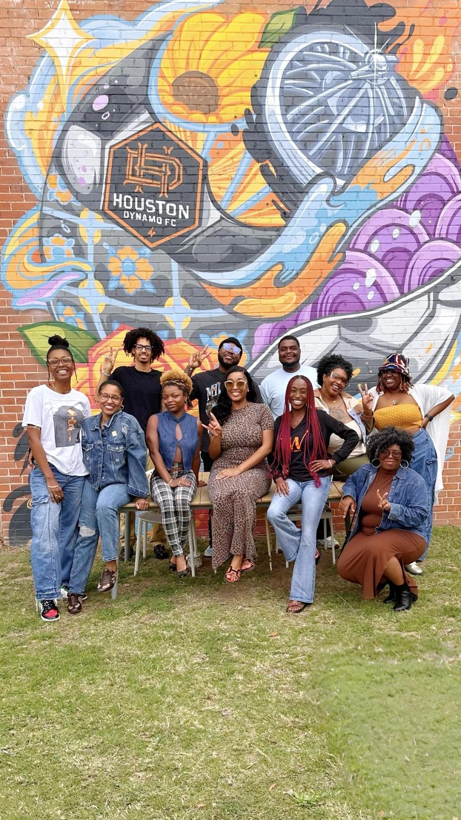 A group of creatives posing in front of a colorful Houston Dynamo FC mural on a brick wall