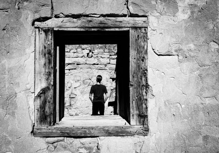 Black-and-white photo of a lone figure framed by a weathered wooden doorway in a cracked stone wall, facing away