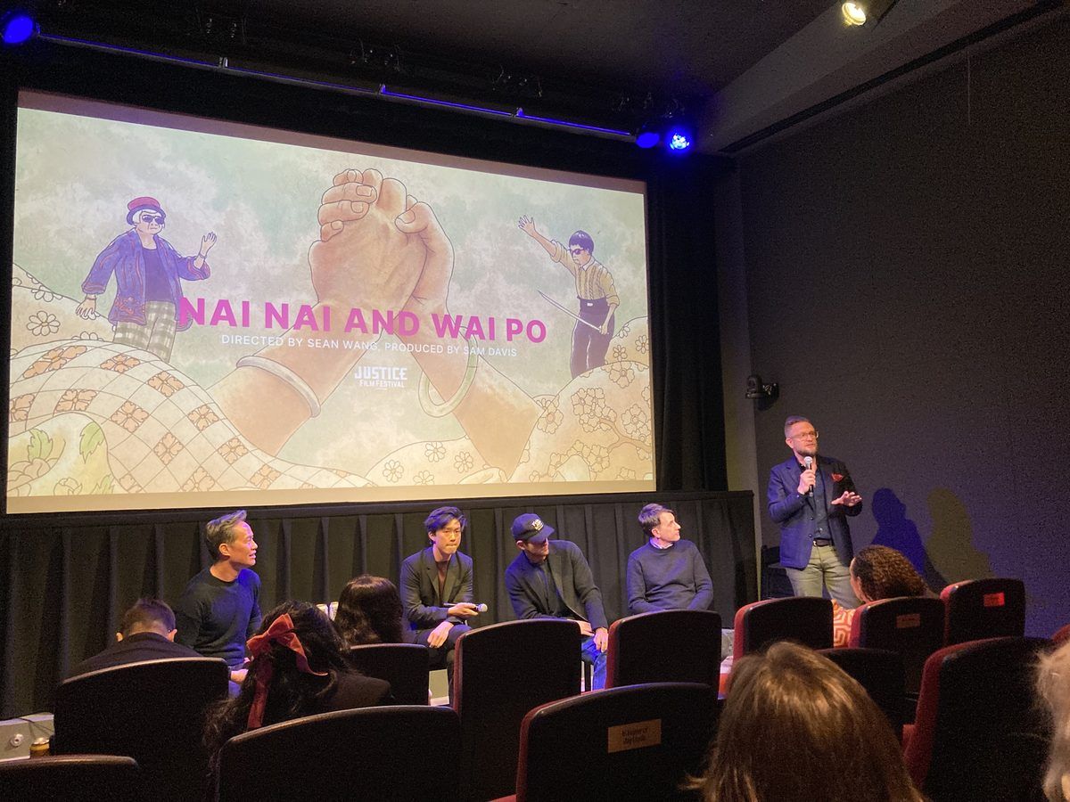 Man introducing a panel of four individuals seated in front of a movie title screen in theater setting