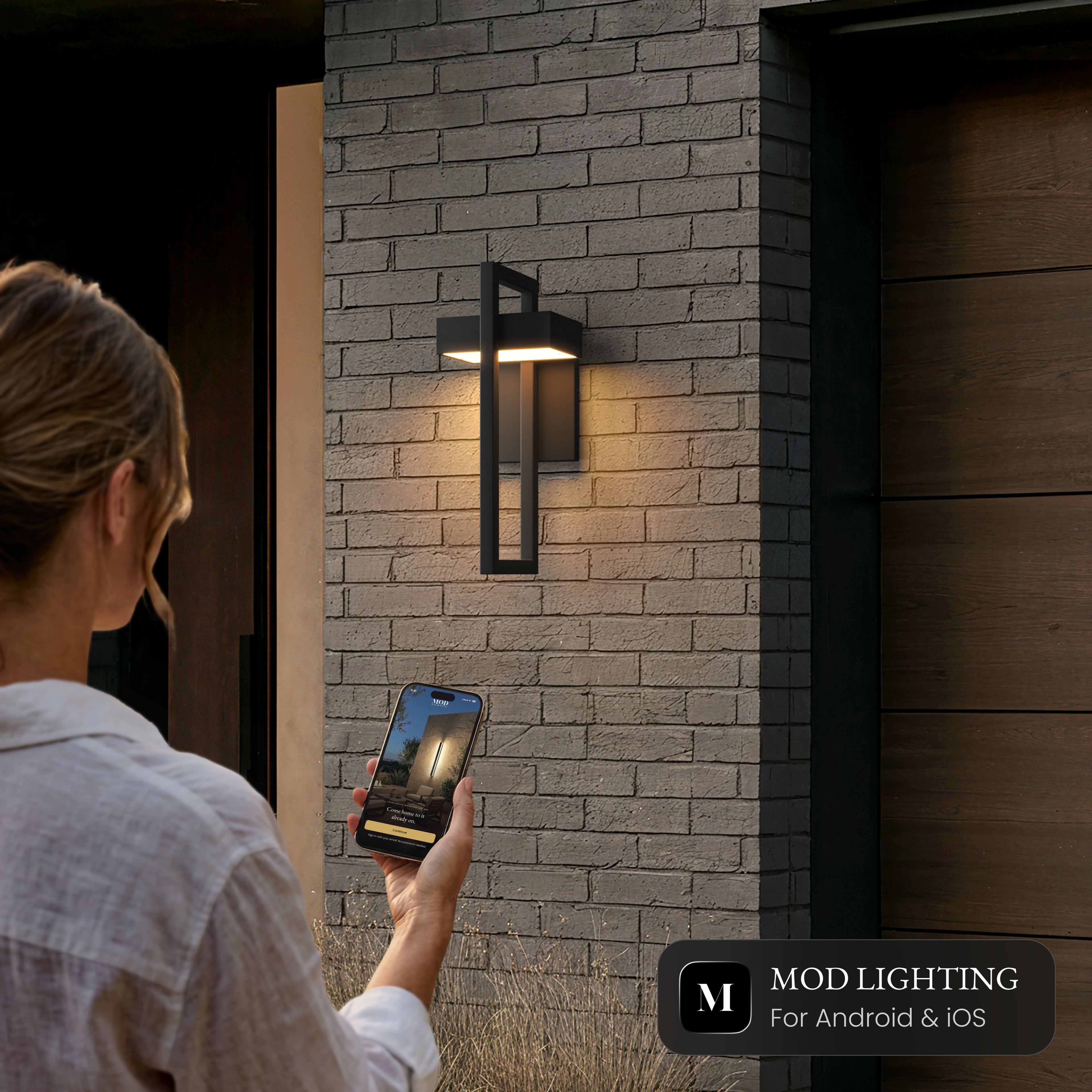 Terri Smart black outdoor sconce on a brick wall, controlled from the MOD LIGHTING app on a phone